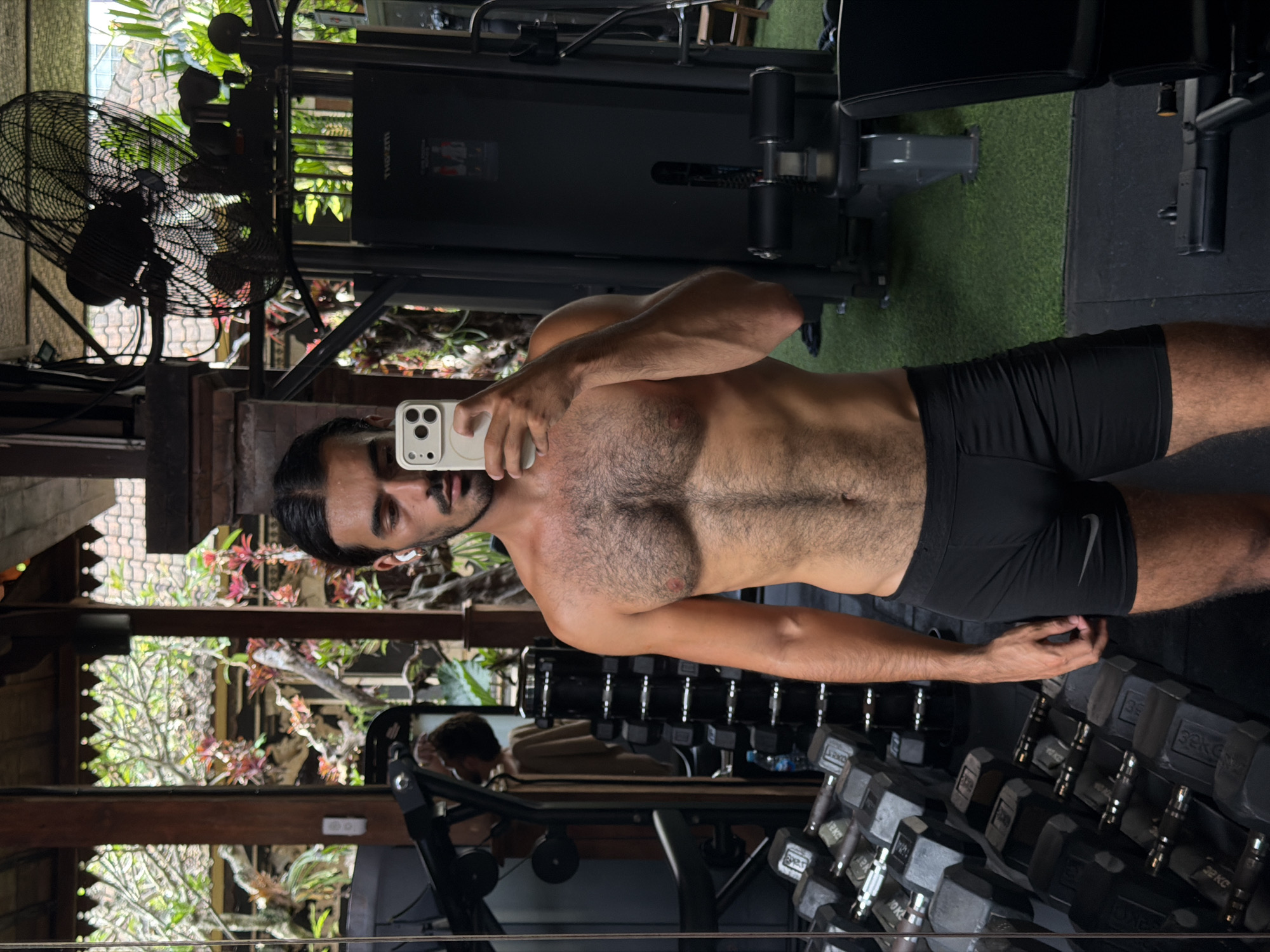 A shirtless man takes a selfie in a gym in North Kuta, Indonesia. He is wearing black athletic shorts with a Nike logo. The gym has weightlifting equipment, including a weight machine, dumbbells on racks, and a fan. Outside the gym, there are tropical plants and trees. A reflection shows another person in the gym.