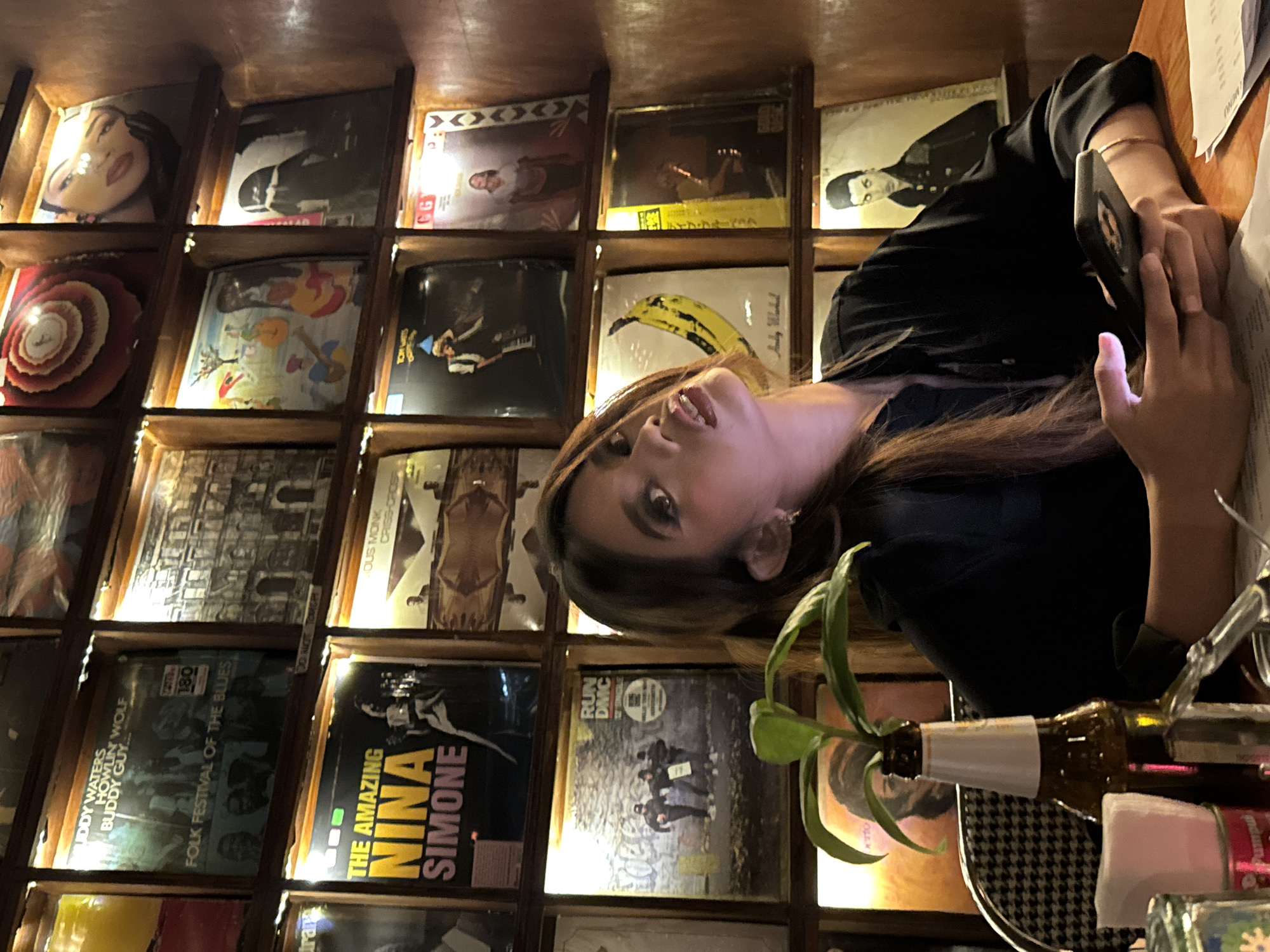 The image shows a young woman with brown hair, looking up and to her right, holding a smartphone in her hands. She is seated at a table with a bottle of beer and some papers. Behind her, a wall is covered with vinyl records displayed in wooden shelves, many of which are illuminated by string lights. The records include artists such as Nina Simone, Run DMC, and Buddy Waters. The background appears to be an indoor establishment with a rustic and vintage ambiance. The setting is likely a bar or restaurant in Davao City, Philippines, given the user's request.