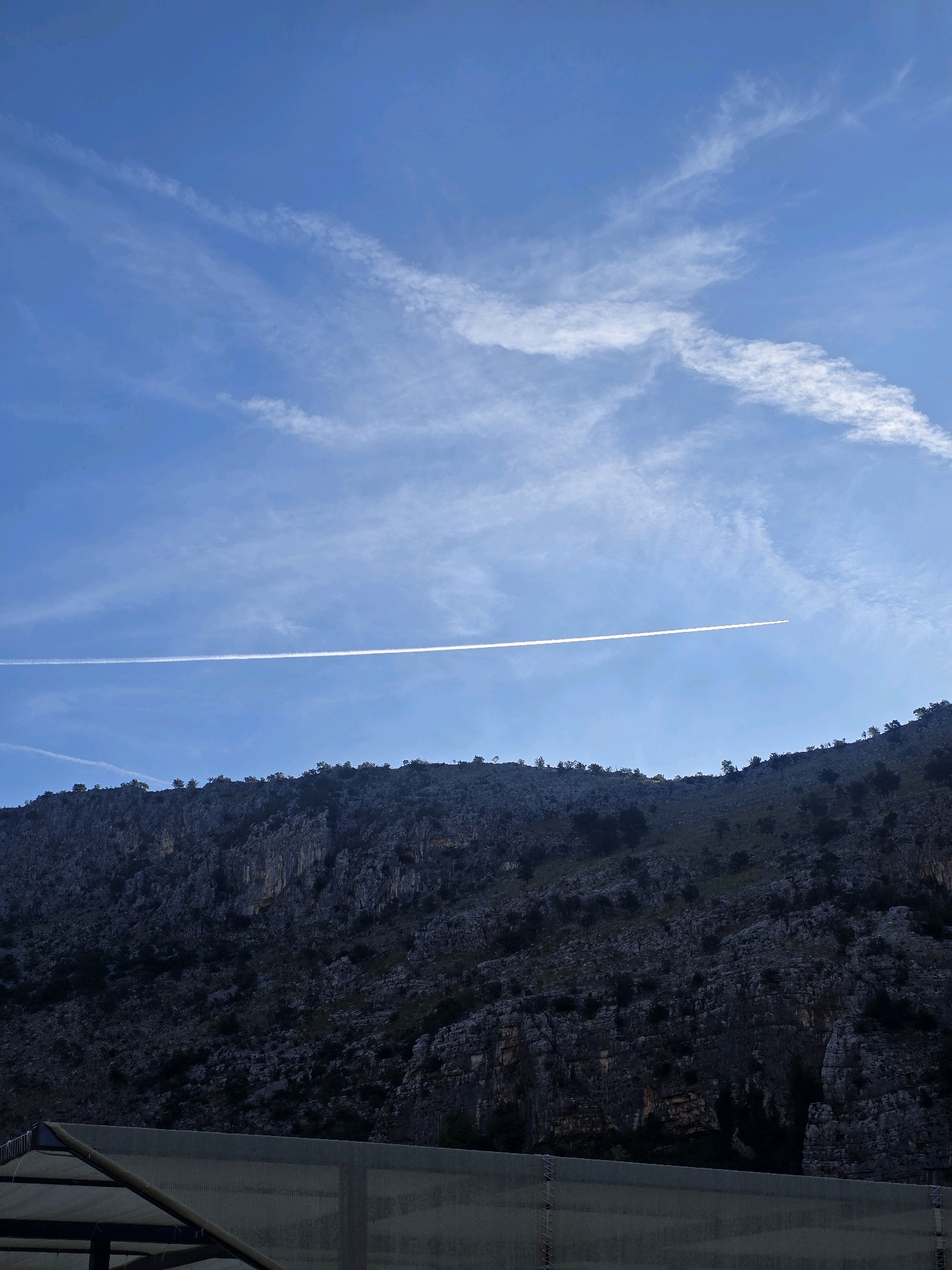 An airplane contrail streaks horizontally across a vibrant blue sky, dappled with wispy white clouds. Below, a rugged, dark-hued mountain range rises, its rocky face textured and dotted with sparse trees. In the foreground, a section of a greenhouse structure, made of metal framing and translucent sheeting, is visible. The overall scene suggests a natural landscape with a touch of human intervention, likely on the outskirts of Podgorica, Montenegro, given the geological formations typical of the region.