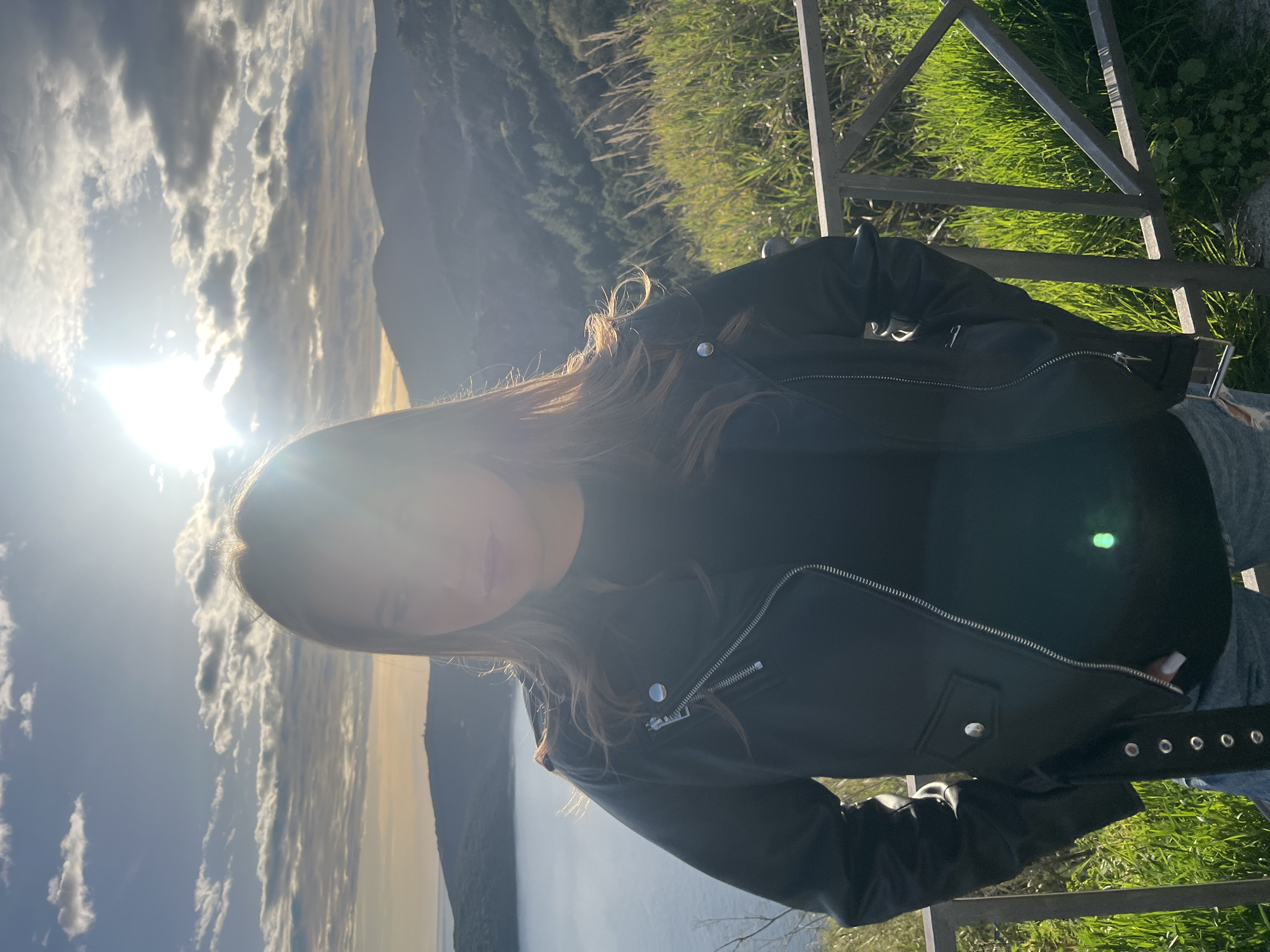 The image shows a person wearing a black leather jacket standing outside. The background features mountains, clouds, and what appears to be a body of water. There's also a railing and green grass present in the foreground. The specific location within the city of Prijevor, Montenegro cannot be determined from the image.