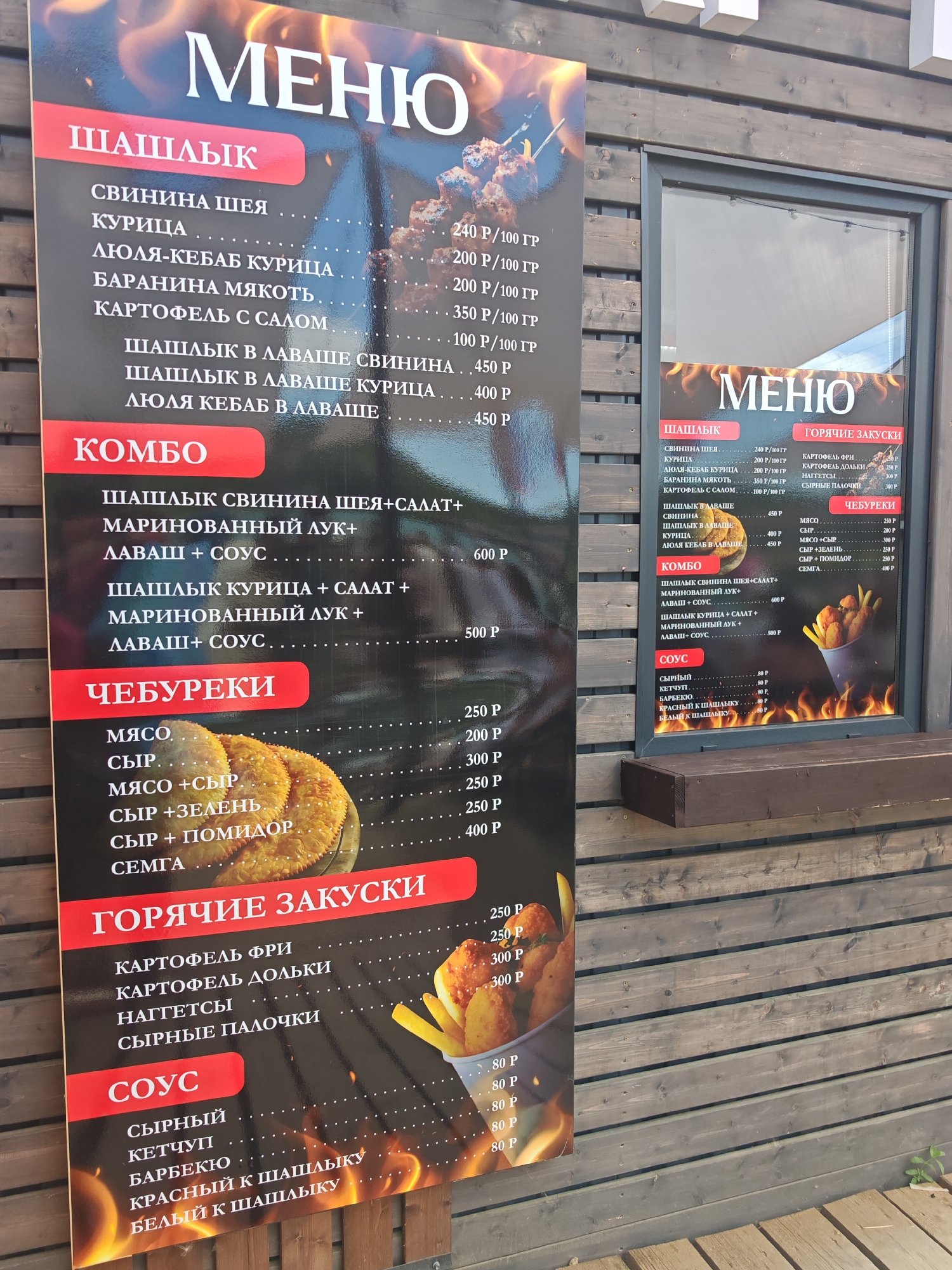 The image shows a menu board outside a building that appears to be a food establishment. The menu board is black with large red lettering and white text listing various food items. The establishment is located in the city of Sukko, Russia. The menu includes sections for "Шашлык" (Shashlik/Skewers), "Комбо" (Combo), "Чебуреки" (Chebureki/Fried Pies), "Горячие закуски" (Hot Appetizers), and "Соус" (Sauce). Prices are listed next to most items. There are also illustrative images of food on the menu board. The background consists of a wooden wall and a window. There are no people or animals visible in the image.