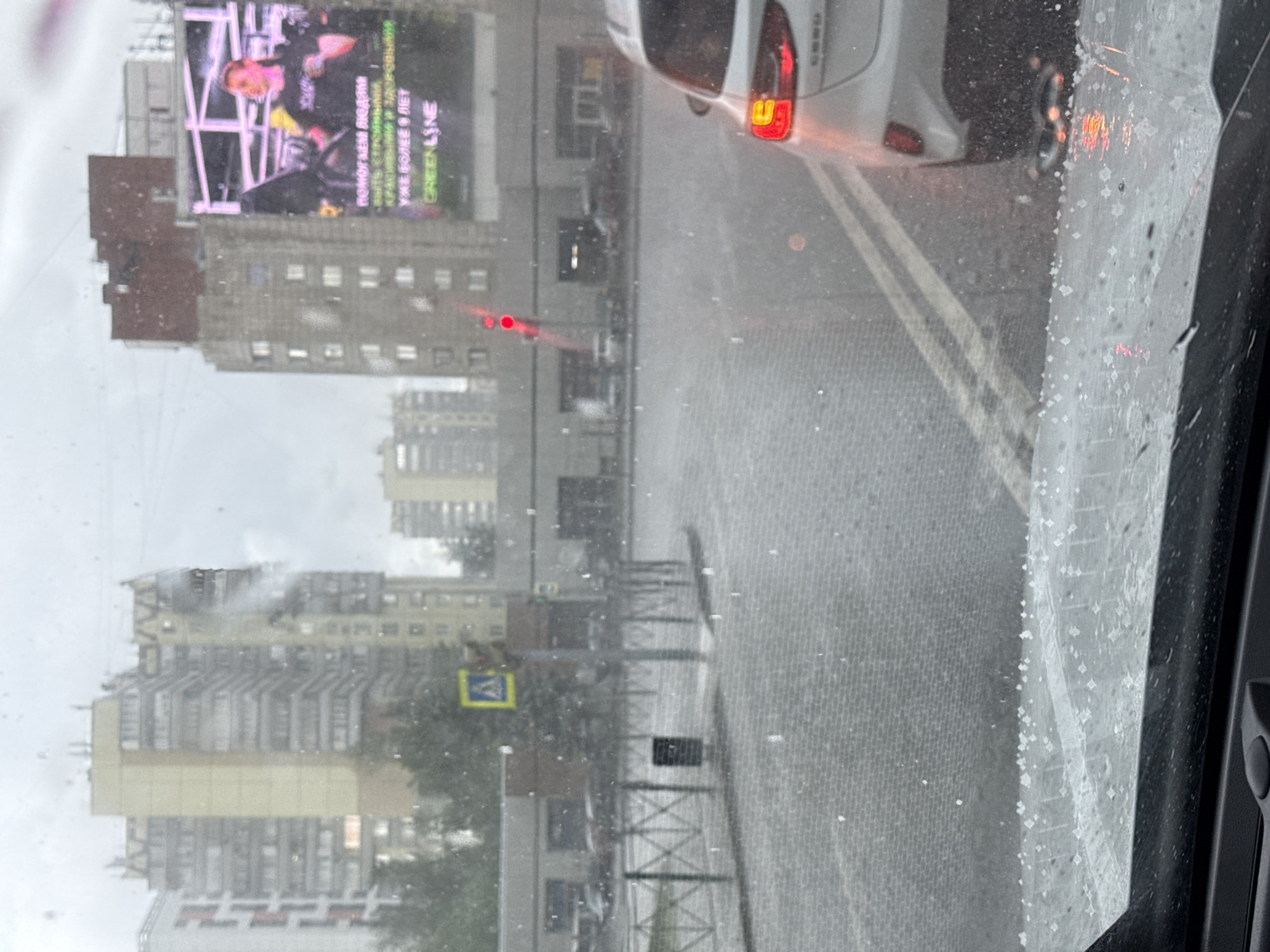 The image depicts a street scene in Perm, Russia, viewed through a rain-splattered car windshield. A white car is in front, brake lights illuminated, on a wet asphalt road with double yellow lines. High-rise buildings dominate the cityscape, including one with a large billboard featuring a person and text in Russian. A pedestrian crossing sign is visible on the sidewalk. Rain is falling heavily, obscuring details of the surroundings. A metal fence lines the sidewalk.