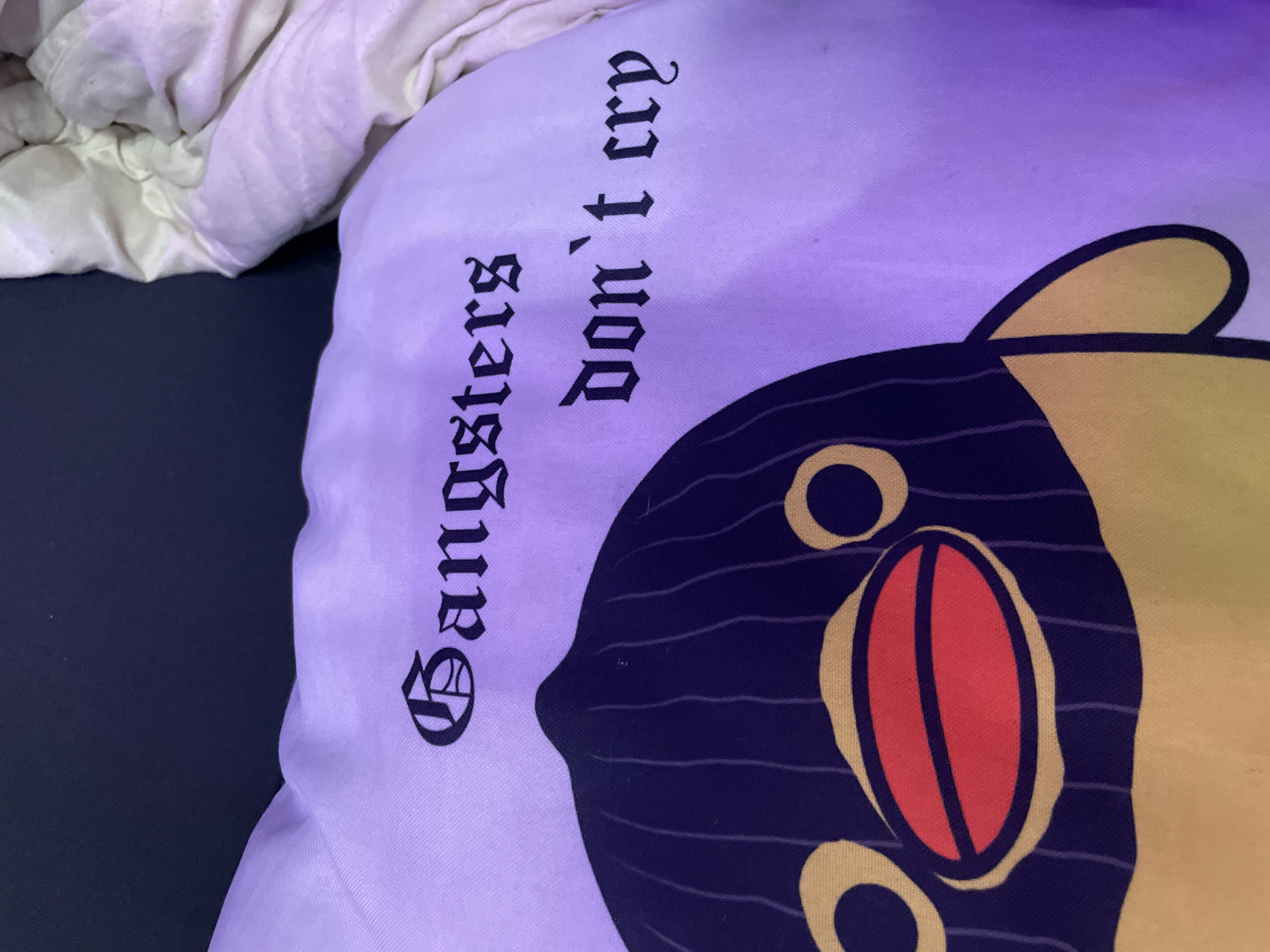 A close-up, overhead shot shows a light purple pillow with a cartoon character print and text. The character has a yellow face with dark blue stripes and a red oval mouth. To the right of the character, the words "Gangsters" and "don't cry" are printed in black gothic-style font, oriented vertically. The pillow rests on a light-colored, crumpled fabric, possibly a blanket or bedsheet. The background is dark, and the scene is illuminated by a purplish light, likely from an LED source. There are no people or specific landmarks visible, therefore the location in Sinyavino, Russia cannot be determined.