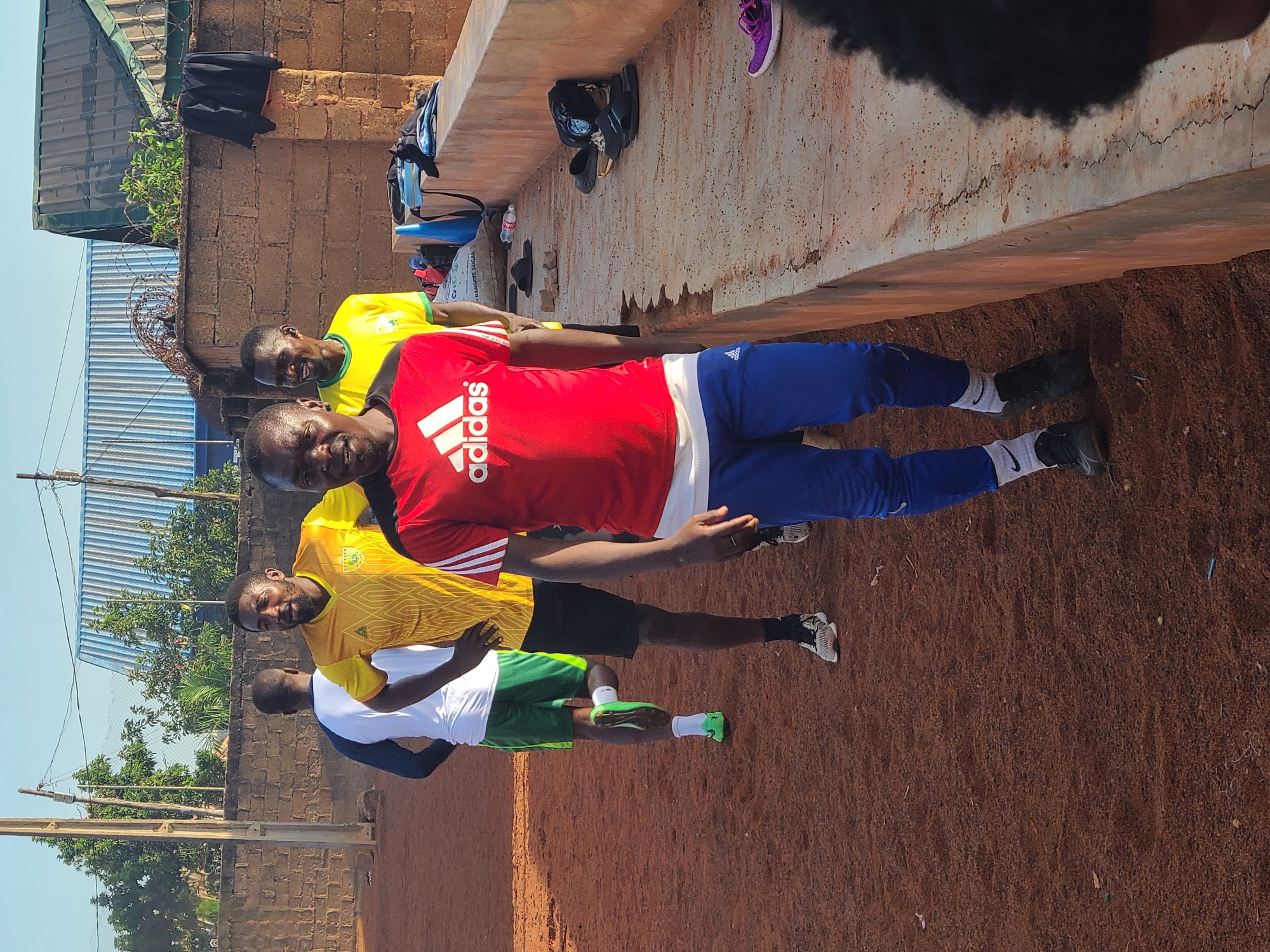 An outdoor, daylight scene captures four adult males gathered on reddish-brown earth, possibly at a sports ground or training area. The central figure, facing the camera, wears a red t-shirt with the white "adidas" logo, blue track pants, and black athletic shoes with white socks bearing a black Nike swoosh. He has a slight smile. To his left, another man in a yellow/gold patterned athletic jersey and black shorts also smiles at the camera. Further left, a third man's head and shoulders are visible in a yellow t-shirt. The fourth man, partially visible on the far left, wears a white long-sleeved shirt, green shorts, and bright green athletic shoes.
In the background, a low concrete wall serves as a makeshift shelf, holding various items: a black jacket, a black backpack, a purple shoe, black sandals, other black shoes, a blue object, a water bottle, and a white bag partially visible with "DANGOTE SUGAR" printed on it. Behind the wall, there are brick structures, a corrugated metal roof, a metal fence with barbed wire, and green foliage. The sky is clear blue, indicating sunny weather. The men appear relaxed, possibly taking a break during a sports activity or training session.