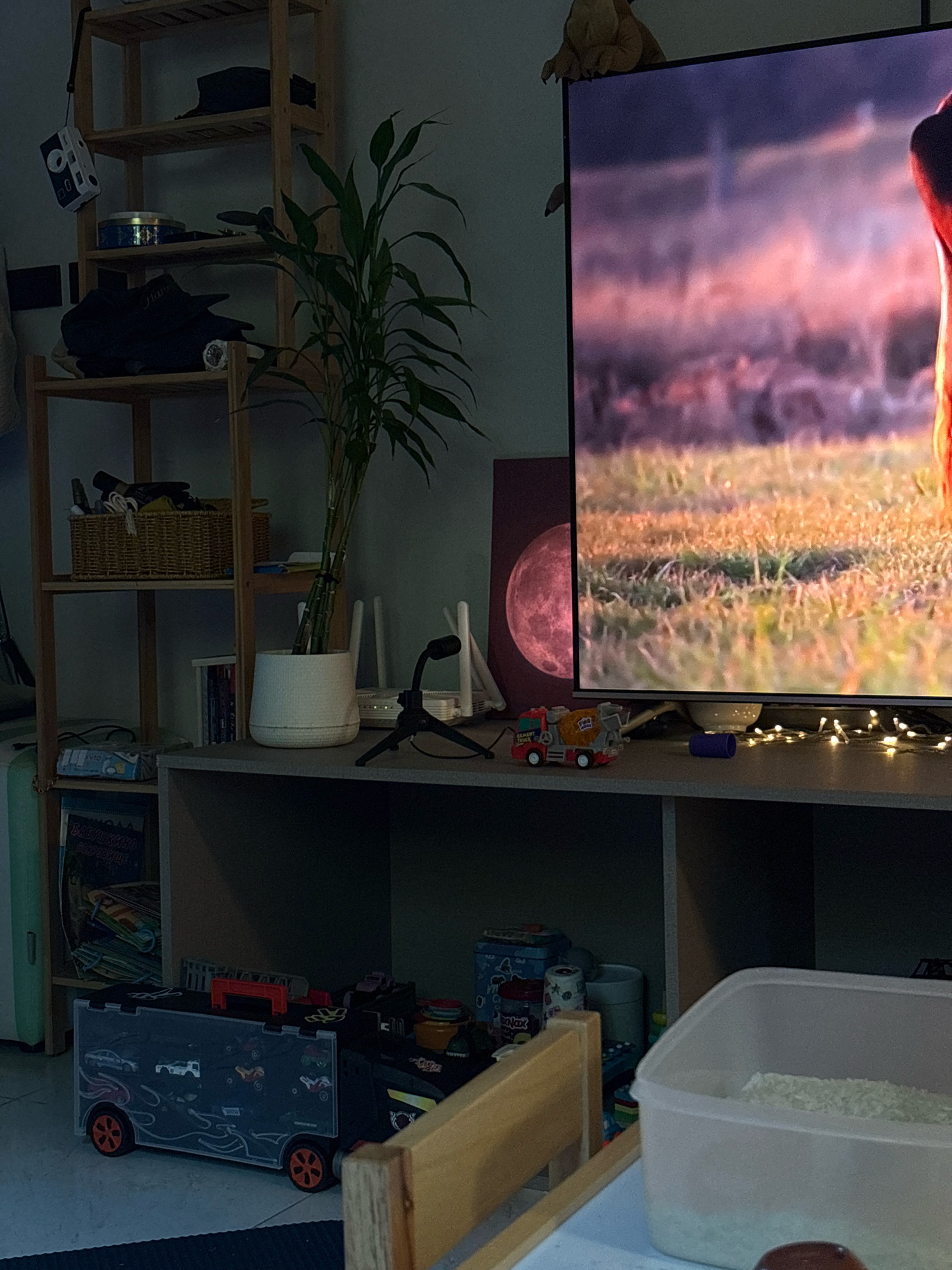 The image depicts a dimly lit indoor scene, likely a living room or play area, situated in Choeng Thale, Thailand. A large television screen displays a vibrant, out-of-focus image of a grassy field bathed in warm light. On the shelf beneath the television, various objects are placed, including a toy cement truck, a purple cylindrical object, and a string of small white lights. To the left, a wooden shelving unit holds a variety of items. The top shelf has a dark object, possibly a hat, and a small retro-style camera. The second shelf contains a woven basket filled with items. Below that, there are stacked items and a potted plant with thin green stalks in a white pot. Further down, a large toy truck carrier with several smaller toy vehicles inside is prominent in the foreground. On the right side, a translucent plastic container filled with white rice is visible. The overall atmosphere is quiet and domestic, with a focus on entertainment and children's toys.