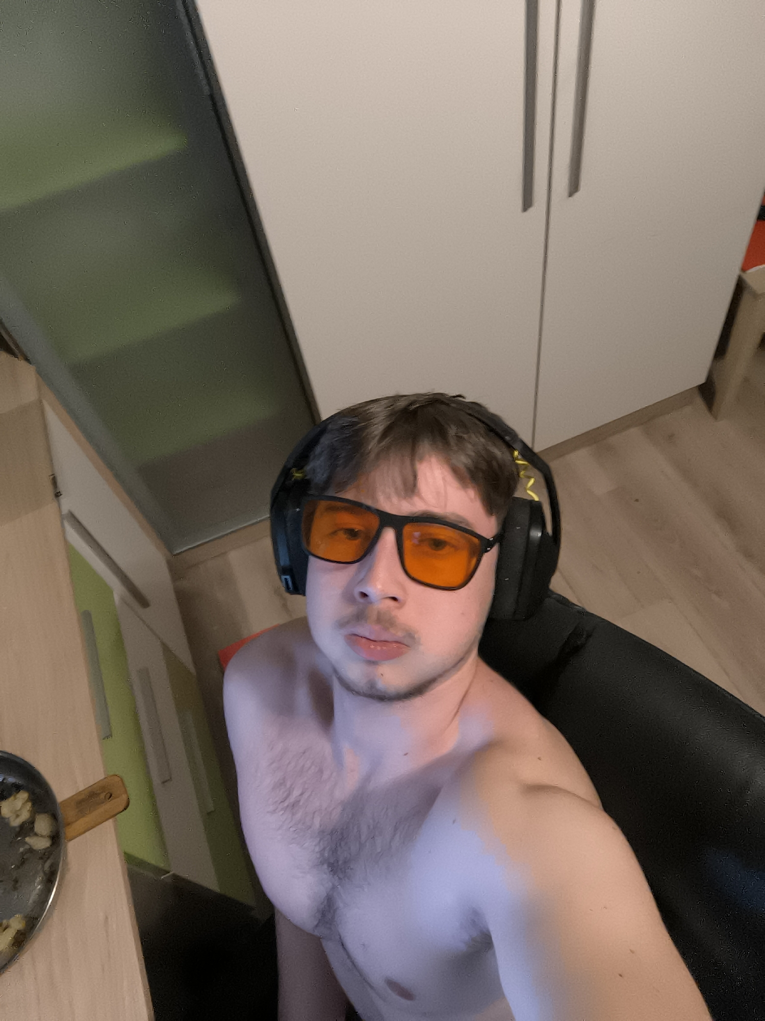 The image shows a shirtless man with brown hair and a slight mustache, wearing black headphones and orange glasses, seated in a black chair. A frying pan with food residue sits on a table to his left. Behind him are light-colored cabinets and a section of the room's floor. There is no information present that reveals the location in the city of Sofia, Bulgaria.