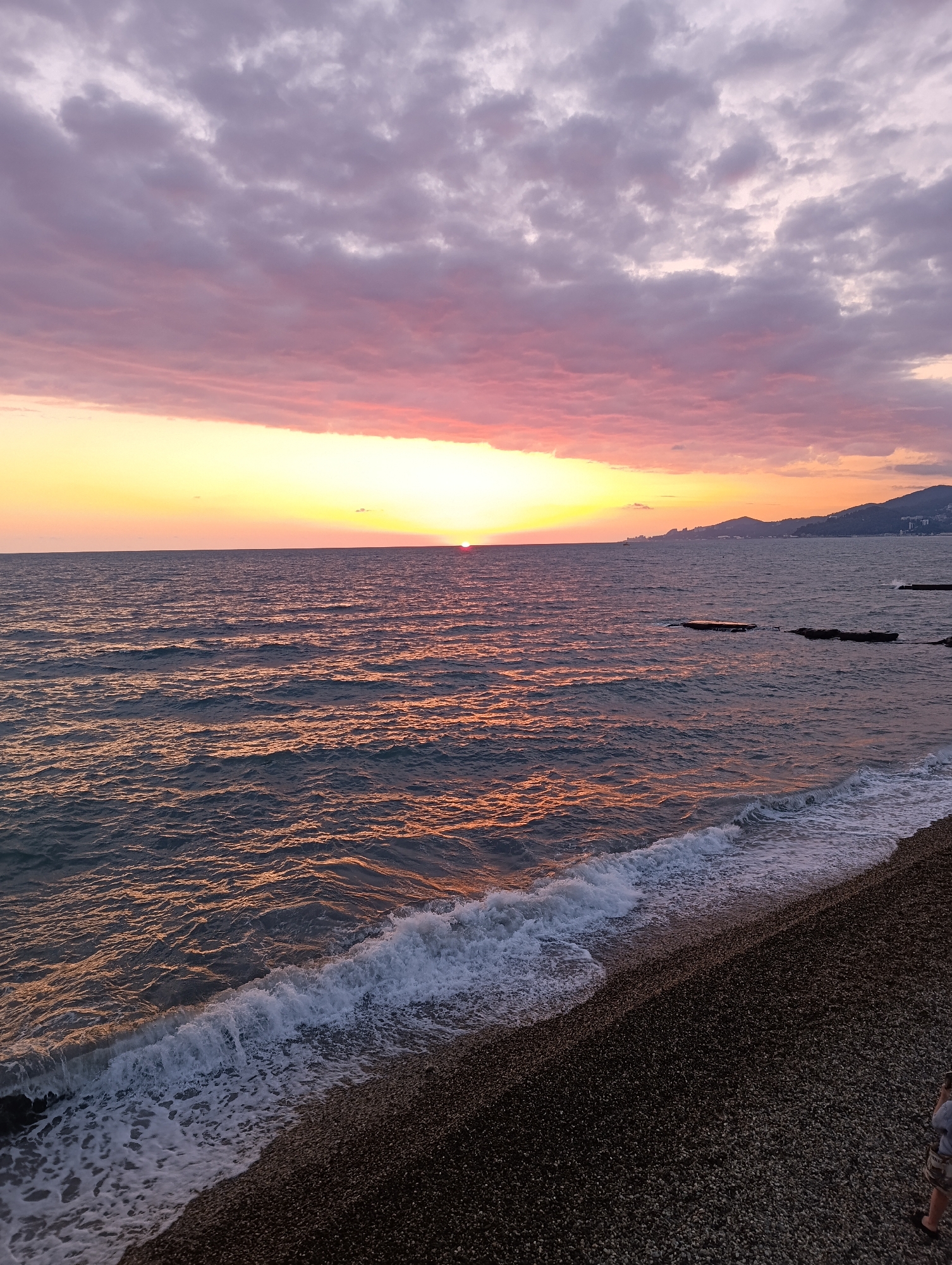 This image captures a serene sunset scene at a pebble beach. The sky is a spectacle of soft pinks and yellows, transitioning into a pale purple as dusk approaches. Wispy clouds are scattered across the horizon, catching the last rays of the sun. The sun itself is a bright, glowing orb, partially dipped below the ocean's edge, casting a warm, golden reflection on the gently rippling water.
The ocean appears calm, with small waves lapping at the shore, creating white foamy lines along the pebble beach. The beach is covered in dark, rounded pebbles, with a few larger rocks visible near the water's edge and some man-made structures, possibly breakwaters or old jetties, jutting out into the sea in the distance. In the background, a hazy silhouette of hills or mountains can be seen, with some buildings dotted along the coastline.
A person is visible in the lower right corner of the image, standing on the beach and looking out towards the sunset. Their presence adds a sense of scale and contemplation to the vastness of the seascape. The overall mood is peaceful and tranquil, evoking the beauty of a day ending by the sea. The time of day is clearly sunset, indicated by the low sun and the colorful sky. The weather appears to be clear and calm, ideal for enjoying the natural display. There is no visible text in the image.