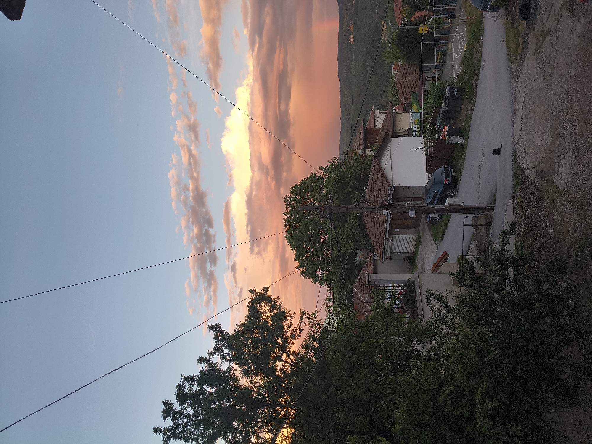 The image displays a picturesque residential street scene in Veliko Tarnovo, Bulgaria, at either sunset or sunrise, characterized by a dramatically colored sky. The perspective is slightly elevated, looking down upon a winding, unpaved road that curves through the middle ground.
Traditional houses with red-tiled roofs are visible on both sides of the street, interspersed with lush green trees and dense foliage. Overhead, utility poles and dark wires cut across the sky. The sky itself is a focal point, featuring elongated clouds illuminated in vibrant shades of orange, pink, and yellow, transitioning into a softer blue further left. A faint, ethereal rainbow-like glow can be observed in the upper right portion of the sky, above a distant, tree-covered hill that defines the horizon.
A small black cat is seen walking in the center of the road, appearing as a solitary figure. A dark-colored car is parked on the right side of the street near a house, with another vehicle partially visible further down. In the middle ground, behind some buildings, a basketball court with a hoop and backboard is visible, along with several dark trash bins. A green and white sign near the court displays partially obscured Cyrillic text, appearing to read "ДЕЛИТА" (DELITA). The scene conveys a sense of tranquility, capturing the blend of daily life with a beautiful natural light spectacle. No people are visible.