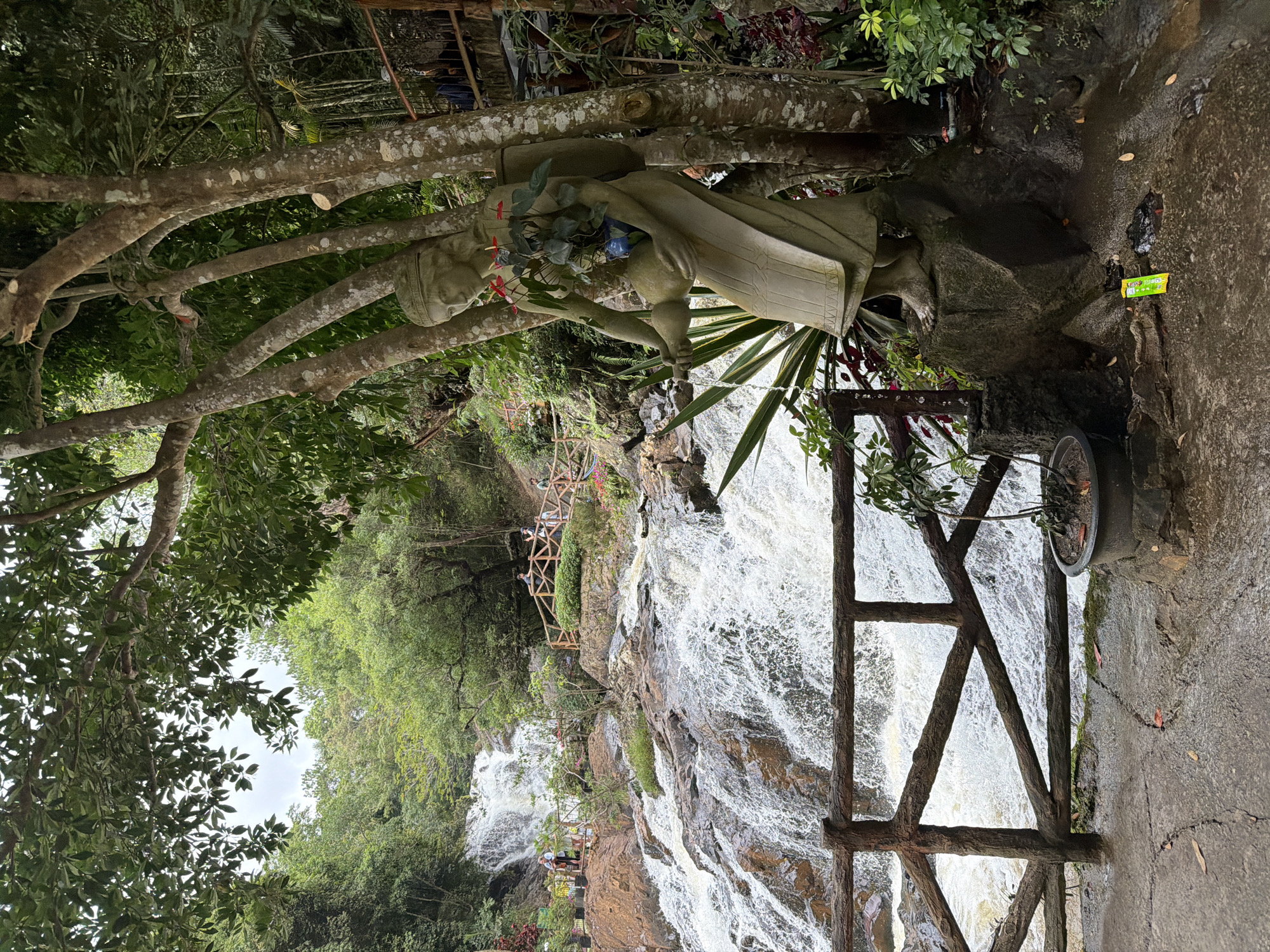 The image captures a scenic view of the Datanla Waterfall in Da Lat, Vietnam. In the foreground, a weathered, stone statue of a woman carrying a pot and a bundle on her back stands beside a thick tree trunk. The statue is positioned on a rocky base, surrounded by lush green foliage and plants. A rustic wooden fence with horizontal rails runs along the left side of the frame, separating the walkway from the cascading water.
The waterfall itself is the central feature of the midground and background. Water rushes down a series of rocky tiers, creating a dynamic and powerful spectacle. A wooden walkway with railings winds its way along the side of the falls, allowing visitors to observe the natural beauty. Several people are visible on the walkway and near the top of the waterfall, enjoying the scenery. The environment is dominated by dense green trees and vegetation, indicating a natural, forested setting. The sky above is overcast, contributing to the diffused lighting of the scene. The overall impression is one of natural beauty and a popular tourist destination.