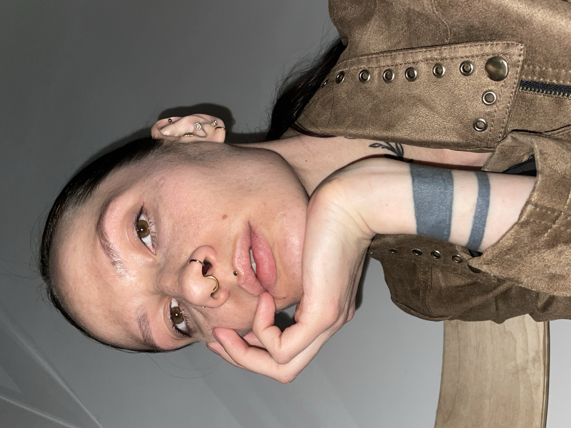 An indoor close-up photograph depicts an adult female, likely situated in Izhevsk, Russia, from the upper torso upwards, looking directly at the camera. Her left hand (viewer's right) is positioned with fingers resting against her chin and jawline.
She has dark hair, brown eyes, and fair skin. Facial piercings include a gold septum ring, a gold hoop in the wearer's right nostril, and a small gold labret stud centered below the lower lip. The visible right ear (wearer's left) features multiple piercings: a gold hoop in the earlobe, a small gold stud, a clear crystal cluster stud, and a small dark red stud in the cartilage.
The female wears a brown, suede-like jacket. The jacket exhibits a front zipper closure and a vertical row of approximately eight metal grommets (eyelets) along the right lapel, secured by a single gold snap button. Tattoos include two thick, parallel dark grey bands encircling the wearer's right wrist and a partial black tattoo depicting a leafy branch on the wearer's right side of the neck/collarbone area.
The background consists of a plain, light grey wall, with a faint diagonal line in the upper left corner, possibly a wall-ceiling intersection. A light-colored wooden surface is partially visible on the far left, behind the subject.