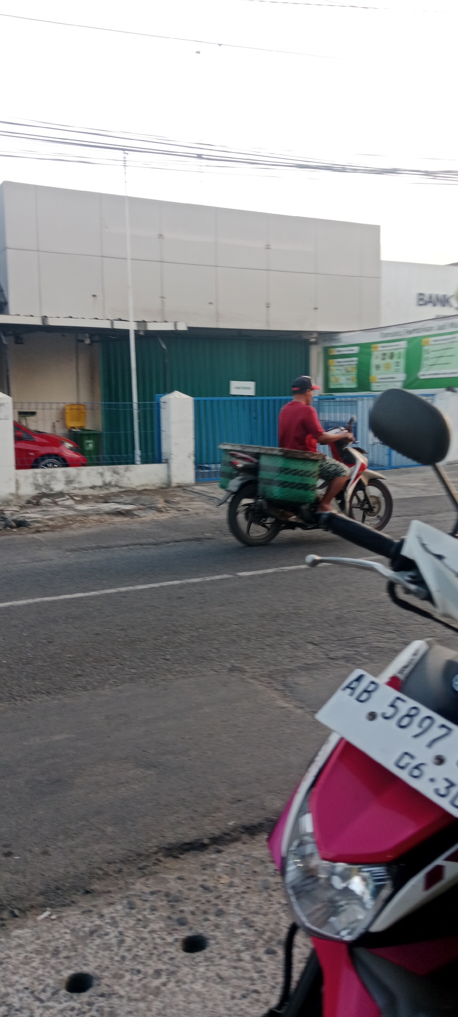 In Maguwoharjo, Indonesia, an outdoor scene shows a person riding a motorcycle on a paved road. The rider, wearing a red shirt and patterned shorts, is looking to the right and has a green container with black stripes attached to the back of the motorcycle. To the left of the road, a building with a white facade and green corrugated metal doors is visible, behind a blue fence. Red and yellow trash bins are present near the building. In the foreground, the front of a pink and white motorcycle is partially visible, with its license plate reading "AB. 5897 G6 .30". The sky is overcast.