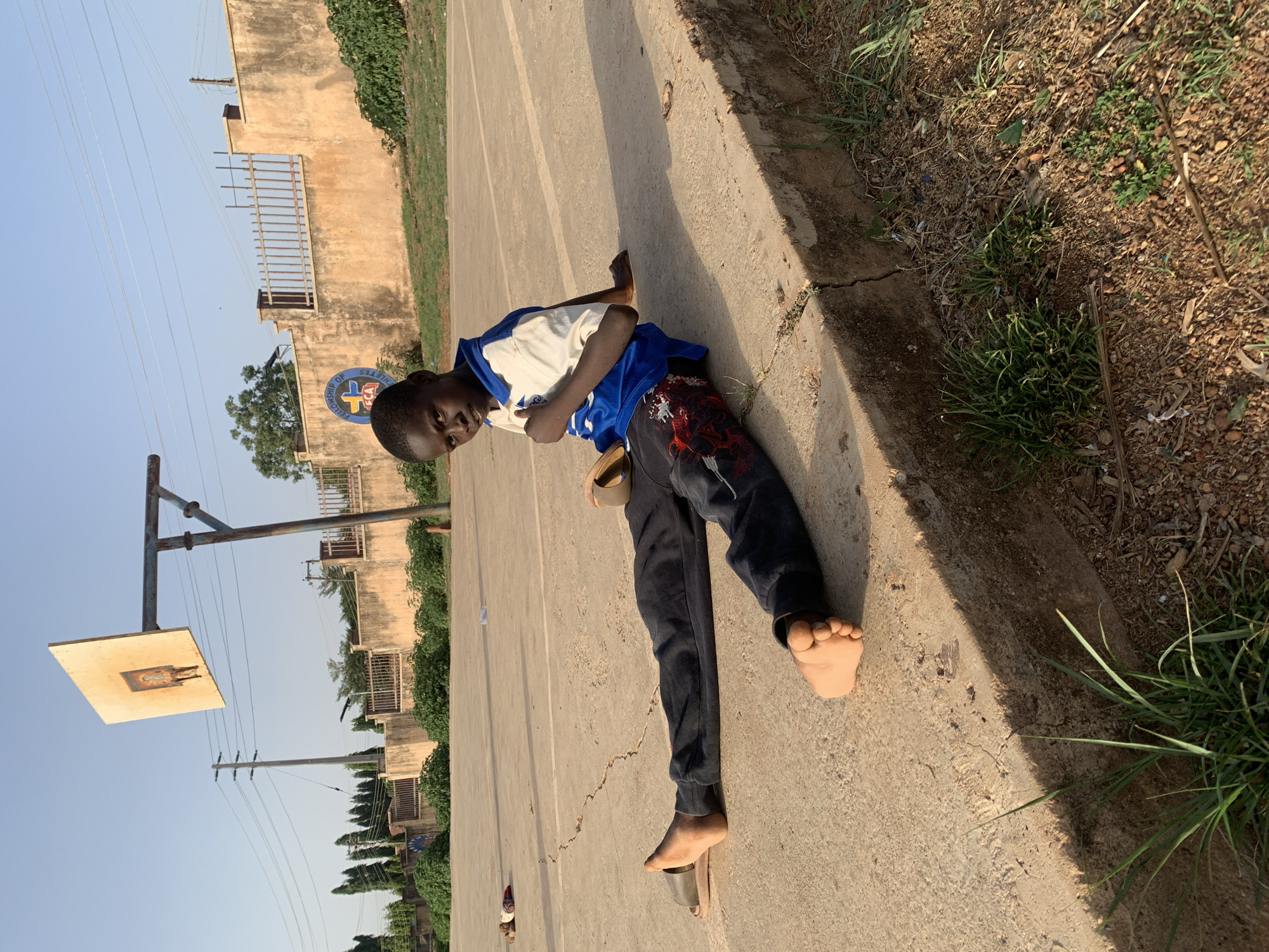 A young Black man lies on his side on a concrete surface, possibly a basketball court, with his head near a basketball hoop in Gura Top, Nigeria. He wears a white t-shirt with blue accents, dark pants, and sandals, with one foot bare. His face is turned towards the camera. The basketball hoop is visibly rusty. A building stands in the background with gated windows. Adjacent to the concrete surface is a strip of dry, grassy area. The sky is pale blue.