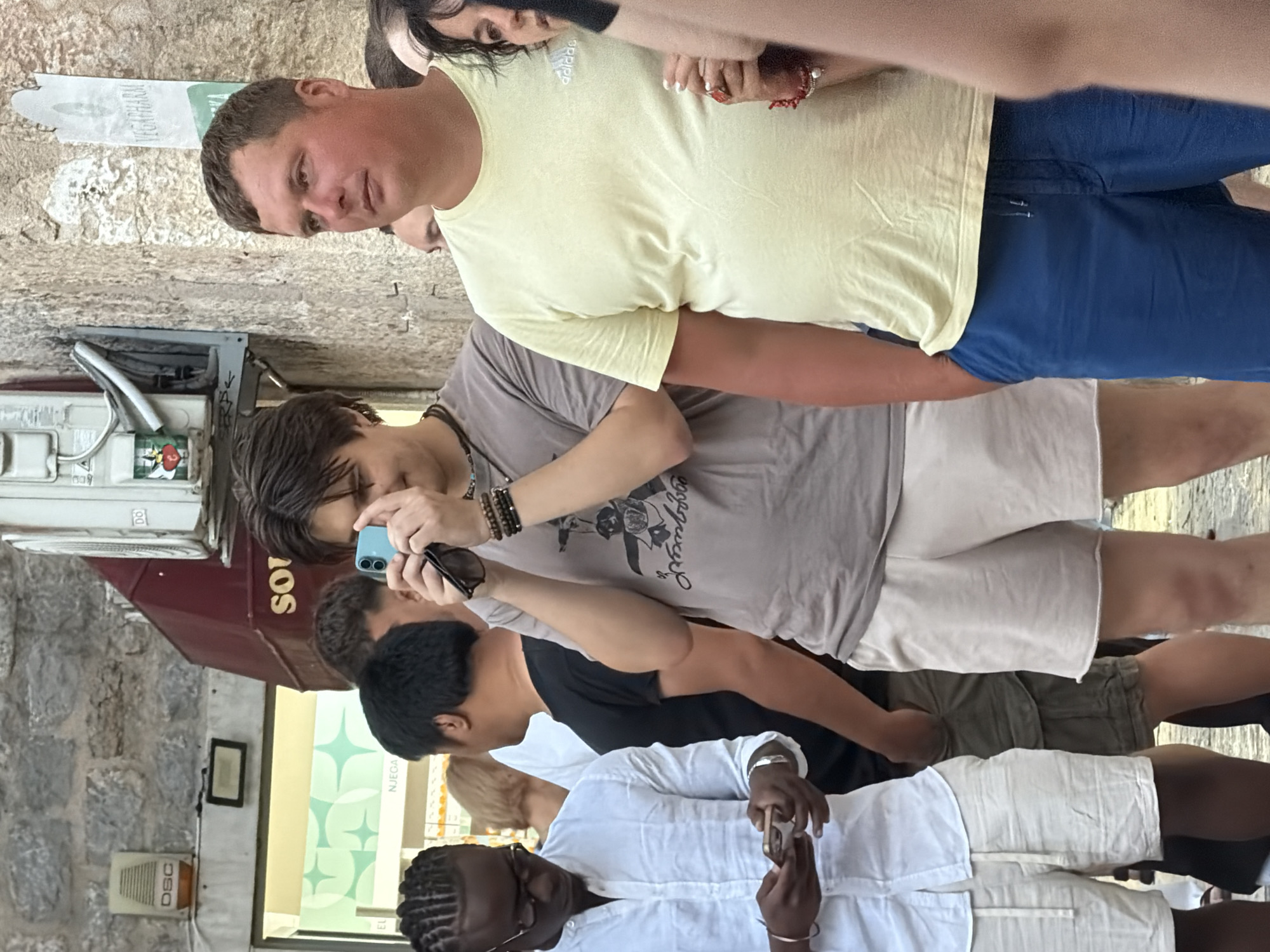 The image depicts a street scene in Budva, Montenegro. In the foreground, a man in a light yellow t-shirt and blue shorts stands to the right, with his hands in his pockets. To his left, a young man in a grey t-shirt and beige shorts is holding a blue smartphone to his face, appearing to take a photo. A man in a white linen shirt and light-colored shorts is on the far left, looking down at his phone. Behind them, other people are visible, along with the facade of a building with a red awning and an air conditioning unit. The ground appears to be paved stone. The overall atmosphere suggests a casual urban setting, likely a street or a plaza.