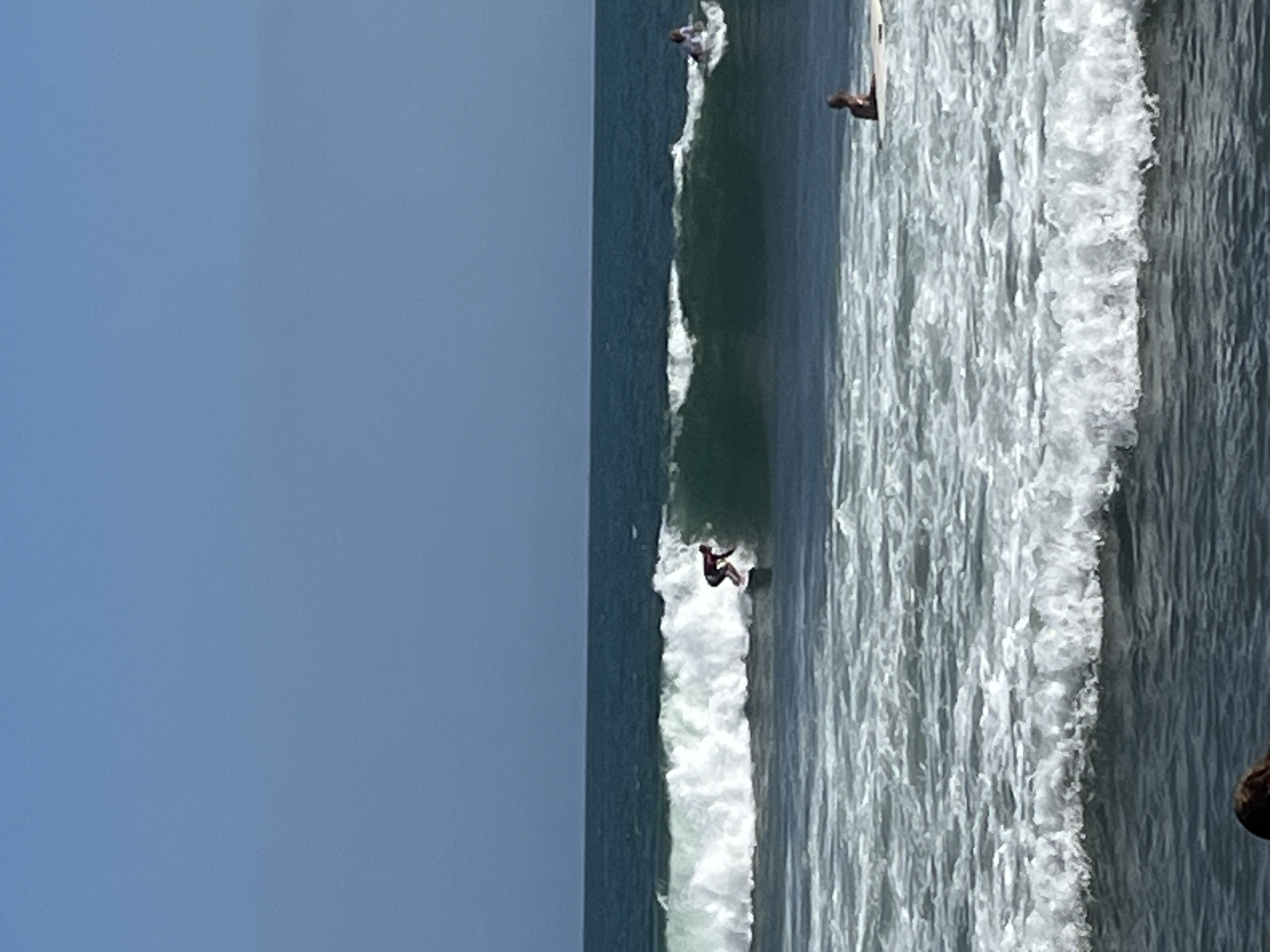 The image shows a coastal scene with surfers in the ocean. In the foreground, there are white, frothy waves. Further out, a larger wave is breaking with a surfer in the process of riding it. This surfer is wearing dark clothing. In the background, another surfer is visible on a wave to the right. The ocean is a deep blue-green color, meeting a clear, pale blue sky. The location is likely a beach in North Kuta, Indonesia, known for its surfing spots.
