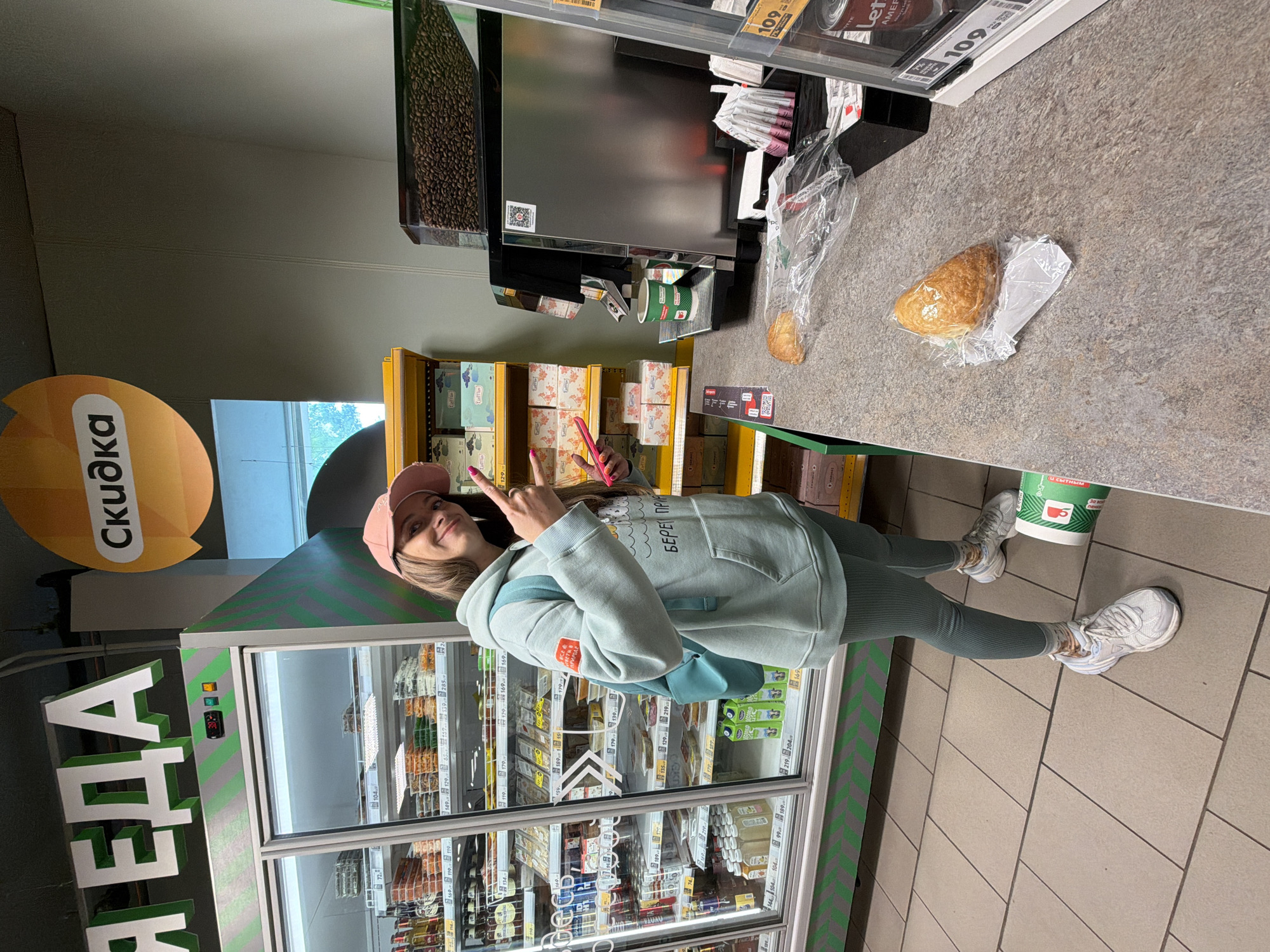 A young woman stands in what appears to be a small shop or convenience store in Birobidzhan, Russia. She is wearing a pink cap, a light blue hoodie, matching leggings, and white sneakers. She is making a peace sign with her fingers while holding a pink object, possibly a phone, in her other hand. Behind her is a refrigerator case filled with various food and beverage items, with the word "ЕДА" (which translates to "FOOD") prominently displayed in green Cyrillic letters. The sign "Скидка" (which translates to "Discount") is visible on a yellow circle. To her right, there is a counter with coffee beans, pastries in plastic bags, and coffee cups. Product prices, such as "109," are visible on shelves near the counter. The environment is well-lit with a tiled floor.