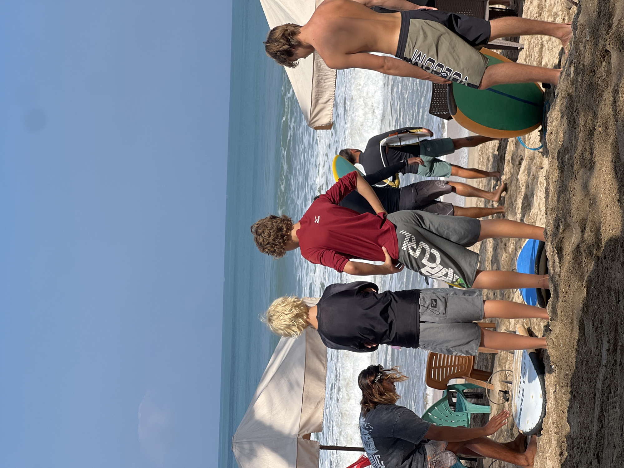 The image captures a sunny day on Kuta Beach, Indonesia, where a group of individuals are gathered on the sand near the ocean. The turquoise water of the Indian Ocean stretches to the horizon under a clear blue sky. Several surfboards are visible on the beach, suggesting surfing activities or lessons are underway. Two large white beach umbrellas provide shade.
In the foreground, two young men face away from the camera, looking towards the water. The one on the left has blonde hair and wears a black long-sleeved shirt and grey shorts. The other, in the center, has curly brown hair and is dressed in a red long-sleeved shirt and grey shorts with a white pattern. To the left, a person with long brown hair, wearing a dark t-shirt and shorts, is bending over near a white surfboard. Further back, two more individuals, also facing away, are partially visible. One wears a black long-sleeved shirt and shorts, holding a surfboard. Another wears a black long-sleeved top with a white stripe on the sleeve and green shorts. The person on the far right is a young man with tan skin, shirtless, wearing olive green and black board shorts, looking out at the ocean. Green and orange inflatable objects are present near the shore. Chairs and other beach furniture are scattered around. The sand is a light brown, typical of tropical beaches. The overall scene conveys a relaxed, outdoor atmosphere.