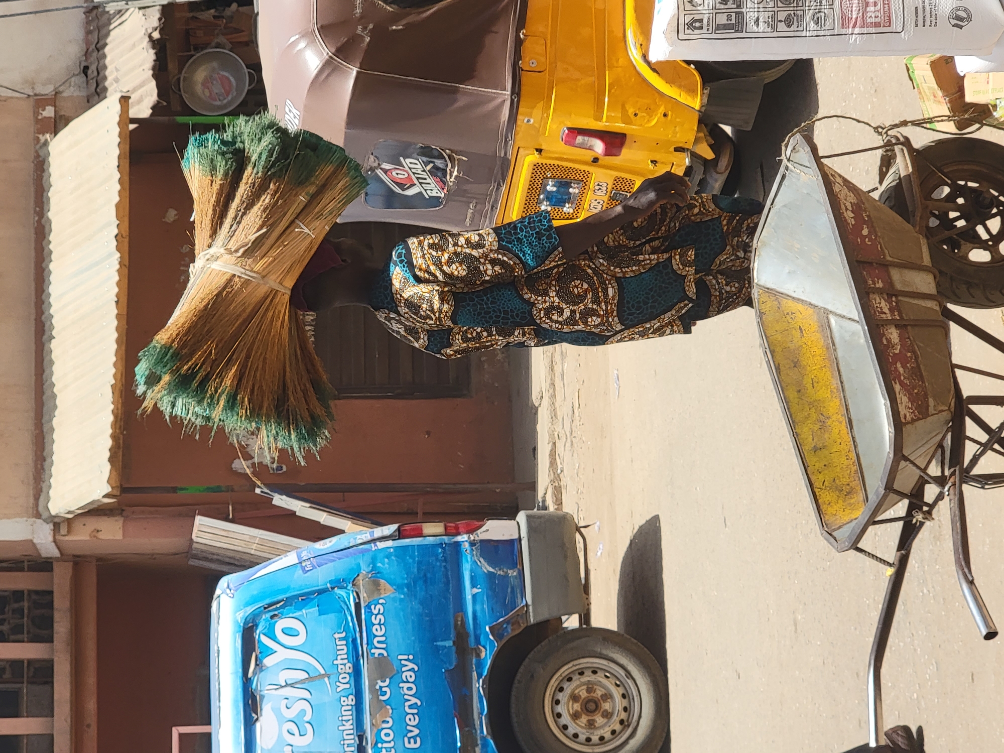 A man in Maiburiji, Nigeria is standing in the street, next to a yellow three-wheeled taxi and a blue van decorated with "Fresh Yo Drinking Yoghurt" advertisements. He wears a patterned blue and gold top. He is surrounded by a bundle of brooms, a white bag, and a yellow and white wheelbarrow attached to a bicycle. The street is concrete and there are buildings visible in the background.