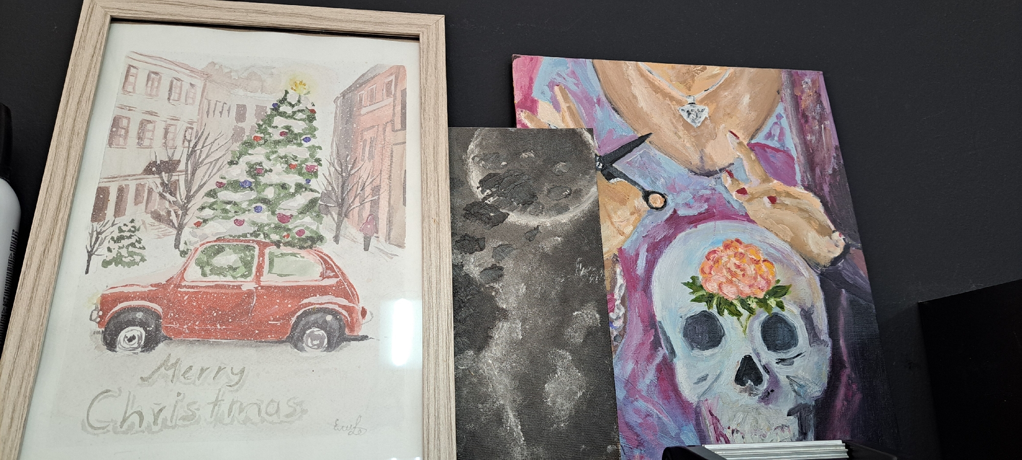 The image displays three framed artworks leaned against a dark wall. The leftmost artwork is a framed watercolor painting of a snowy street scene. It features a red vintage car parked beside a decorated Christmas tree, with snow-covered buildings in the background. The words "Merry Christmas" are written in cursive below the car. The painting is enclosed in a light-colored wooden frame. To the right of this, a narrow, dark abstract painting with a circular motif is visible. The rightmost artwork is a colorful oil painting depicting a stylized skull adorned with a peach-colored flower, set against a backdrop of flowing fabric and hands. Scissors are positioned near the top of this painting. There are no people or distinct landmarks visible in the image that would allow for the determination of its location in Yessentuki, Russia.
