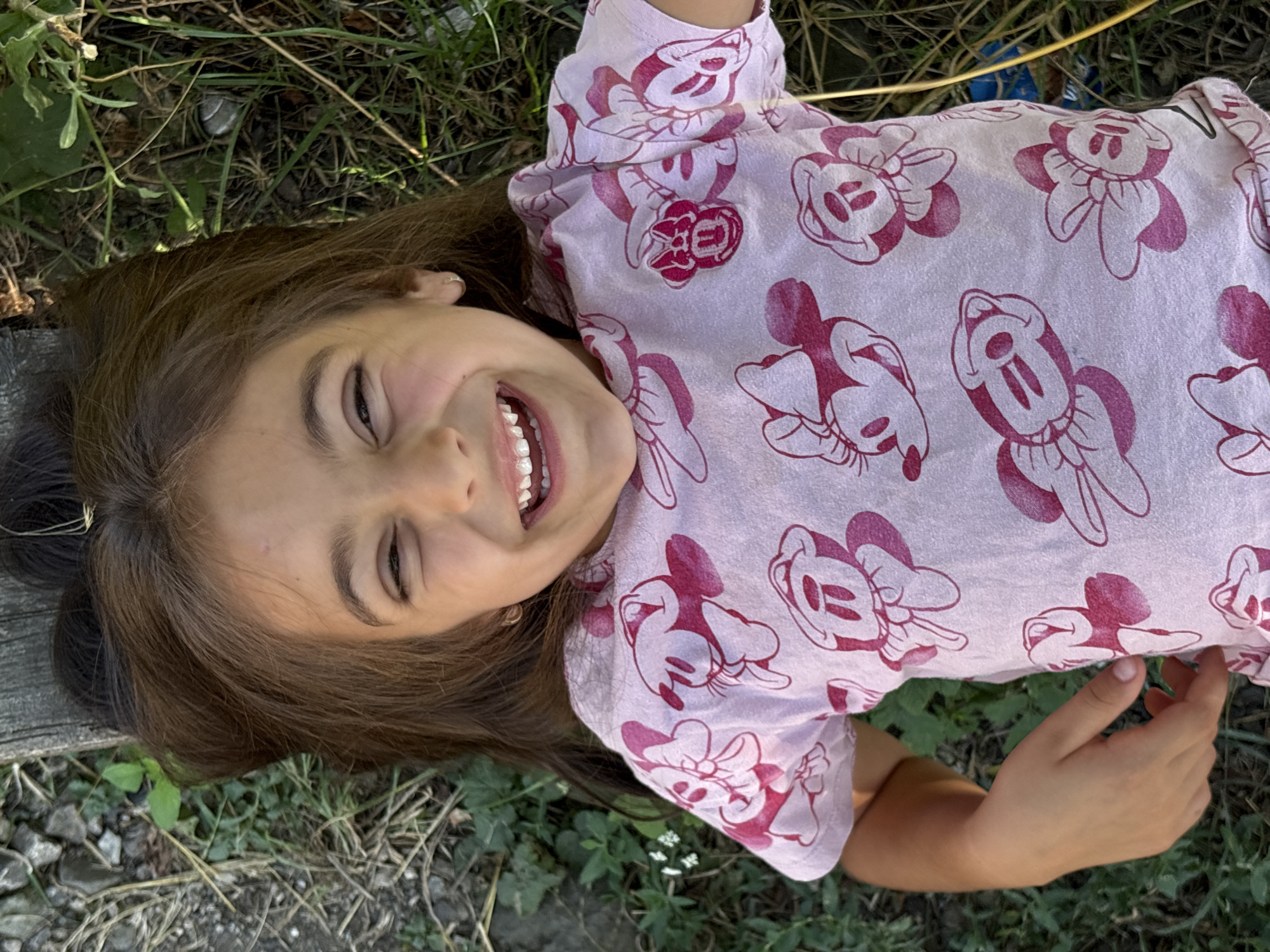 A young girl with long brown hair is lying on her back on the ground in Oreshak, Bulgaria. She is wearing a light pink t-shirt with a repeating pattern of Minnie Mouse characters. Her head is tilted back, and she is looking up and smiling widely, showing her teeth and braces. Her right eye is partially closed. Her left arm is bent, and her hand is resting on her stomach. The ground beneath her is covered with grass, dirt, and some dry leaves, with a wooden plank visible behind her head. The scene is outdoors, likely in a natural or garden setting.