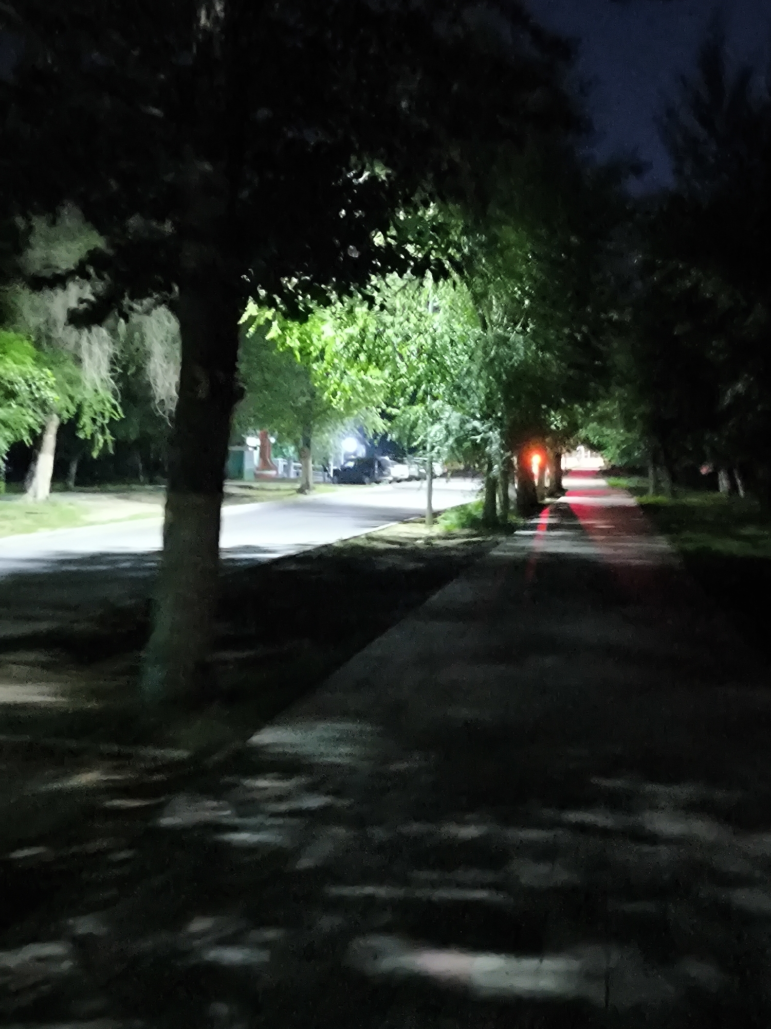 The image depicts a dimly lit pathway lined with trees at night. The pathway appears to be paved, with shadows cast by the trees creating an uneven pattern on the surface. There are some distant lights visible, including what appears to be a red light source, possibly a traffic signal or a building light, casting a red reflection onto the pathway. The trees are dense, and their foliage obscures the sky, which is dark, indicating it is nighttime. In the background, there are hints of a road and some vehicles parked or driving, suggesting an urban or suburban setting. The overall mood is somewhat somber and quiet due to the darkness and the empty pathway. The location is identified as Kostanay, Kazakhstan. There is no visible text in the image. The image captures a common nocturnal scene of a public space, possibly a park or a residential area, illuminated by artificial light sources.