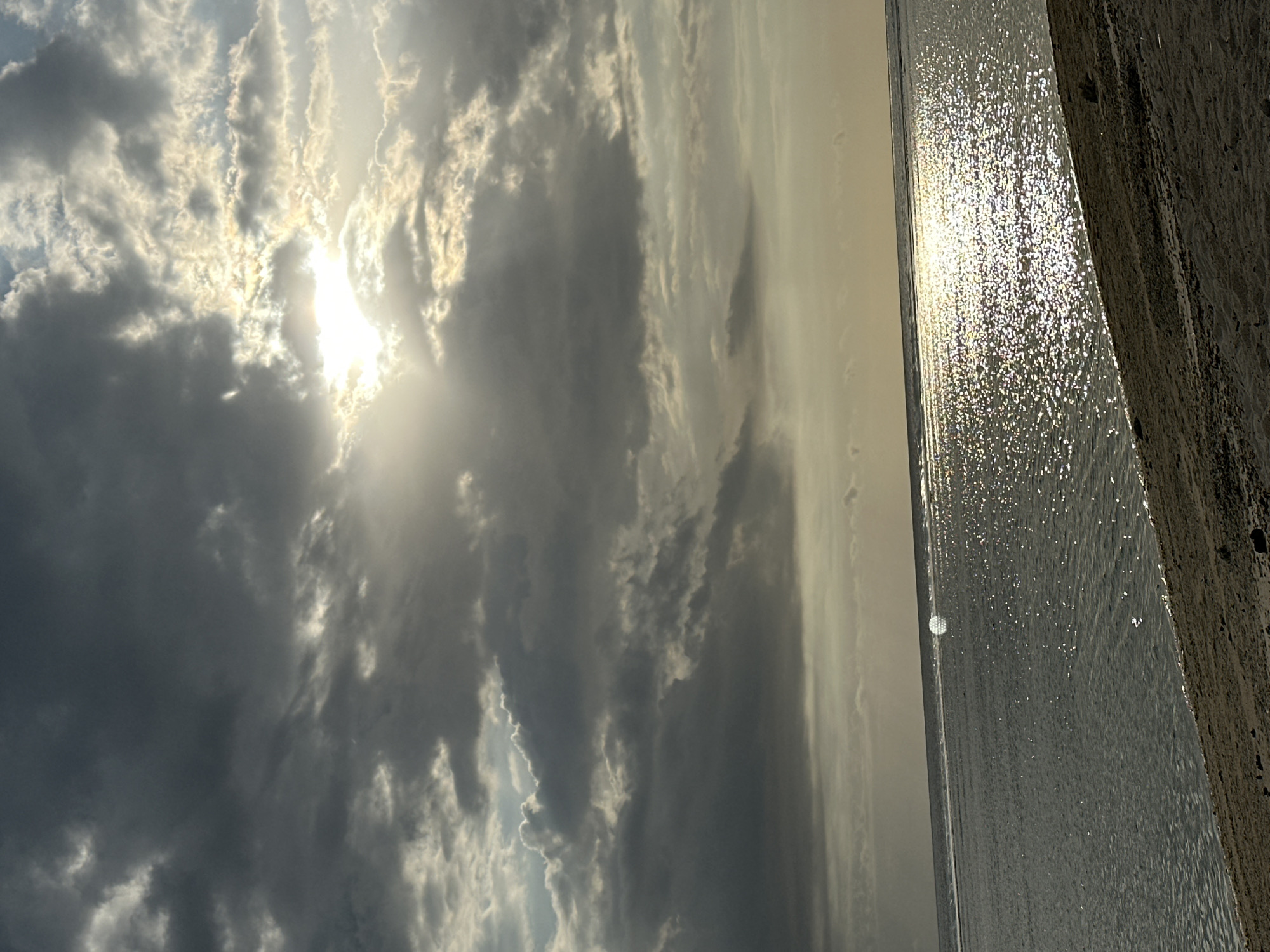 The image shows a beach scene at an unknown location in Indonesia, dominated by a cloudy sky with the sun shining through. The clouds range from light to dark grey, indicating varied density and light penetration. The sea, occupying the right side of the frame, reflects sunlight, creating a shimmering effect. A concrete structure is visible along the right edge, suggesting a seawall or building. The scene lacks specific landmarks or identifying features to pinpoint the exact location within Indonesia. No people are visible.
