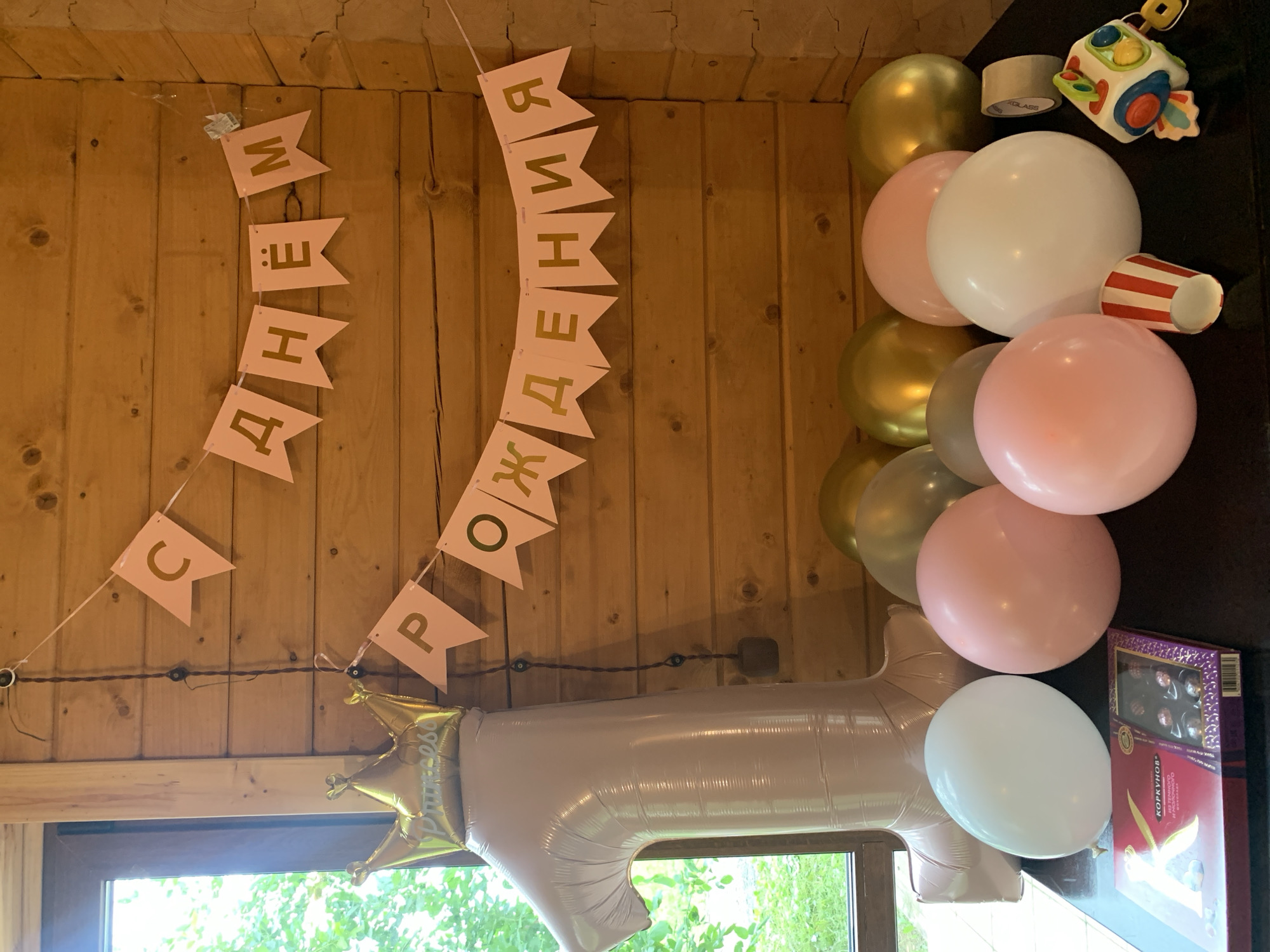 The image depicts a birthday celebration with festive decorations against a wooden wall. A banner with gold letters reads "С ДНЕМ РОЖДЕНИЯ" (Happy Birthday) in Russian, hung vertically on the left side of the frame. Another banner on the lower left has the letters "С", "П", and "Р" visible, likely part of another celebratory message.
Dominating the right side of the image is a cluster of balloons in various colors, including gold, pink, and white. A large, light pink balloon shaped like the number "1" is prominent at the bottom, adorned with a gold crown balloon on its left.
In the background, on a dark surface, are additional party items. These include a small toy, a roll of tape, and a striped paper cup. A box of chocolates, with Russian text that appears to say "КОРКУНОВ", is also visible.
The setting appears to be an indoor space, possibly a room in a wooden house, with natural light filtering in from a window at the bottom, suggesting it is daytime. The overall mood is festive and cheerful, indicative of a birthday party. The location is Chaikovsky, Russia.