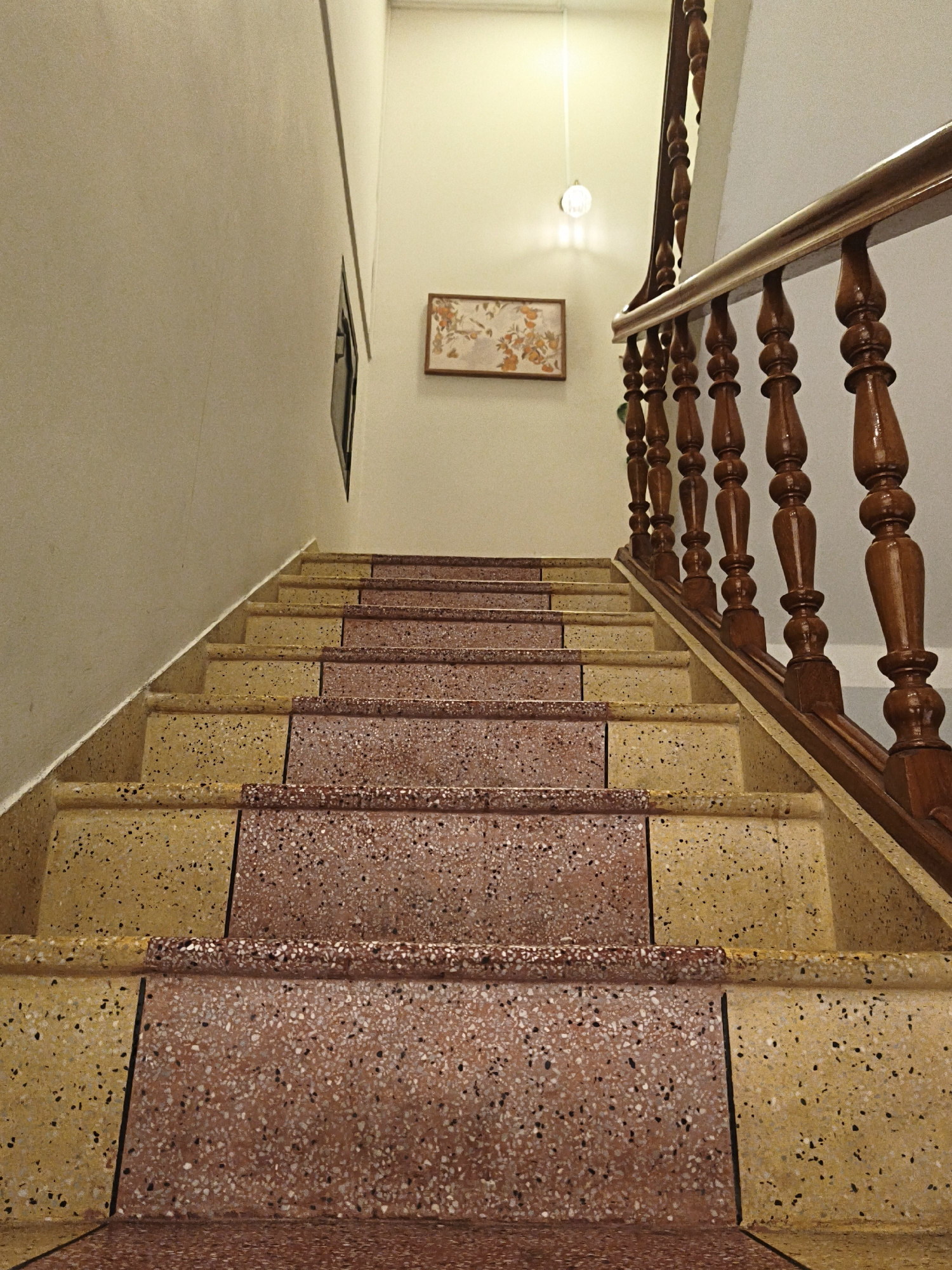 The image shows a view looking up a staircase. The steps are made of terrazzo material, with alternating colors of yellow and reddish-brown. The riser of each step is a darker shade of the same color as the tread. A wooden bannister with turned balusters runs along the right side of the staircase. The wall to the left is a neutral cream color, and on the opposite wall, further up, is a framed artwork depicting flowers and a small, dark-framed window. A light fixture is visible on the ceiling above. The scene appears to be indoors, likely in a residential or commercial building.