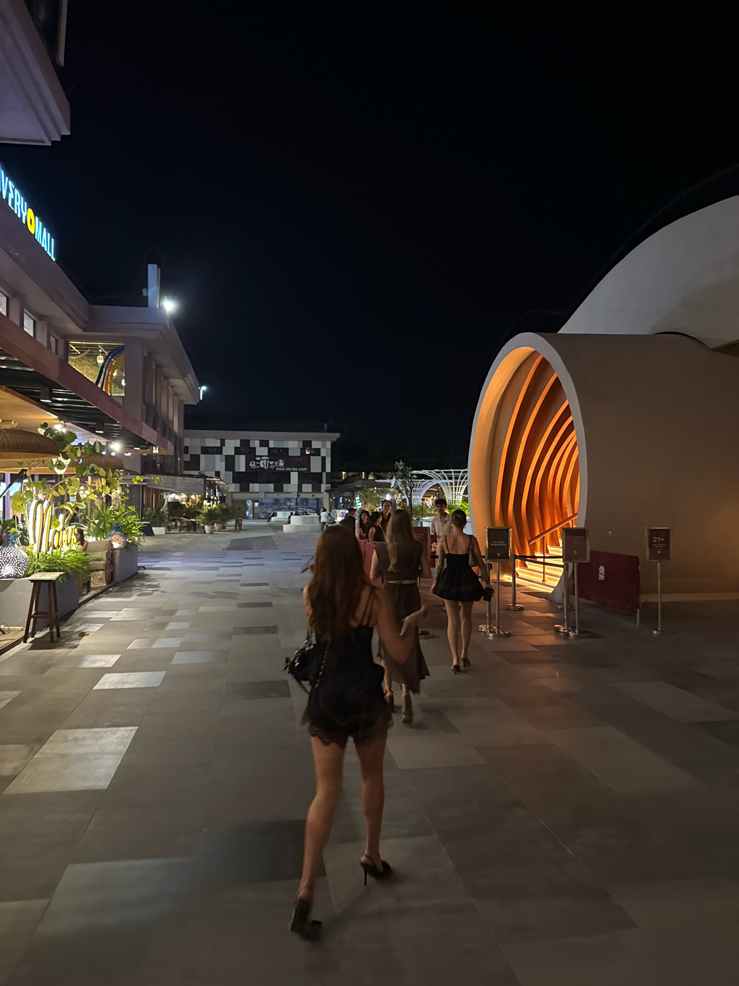 In Kuta, Indonesia, at night, a group of women walk down a tiled pathway. The woman in the foreground, viewed from behind, wears a black lace dress and high heels. She carries a black handbag. To her left, a row of shops with potted plants and illuminated decor lines the pathway. To her right, a large, abstract architectural structure with an arched entrance glows with warm interior light. Several other people are visible further down the pathway, some in the distance near another building with a checkered facade. The sky above is dark and clear.