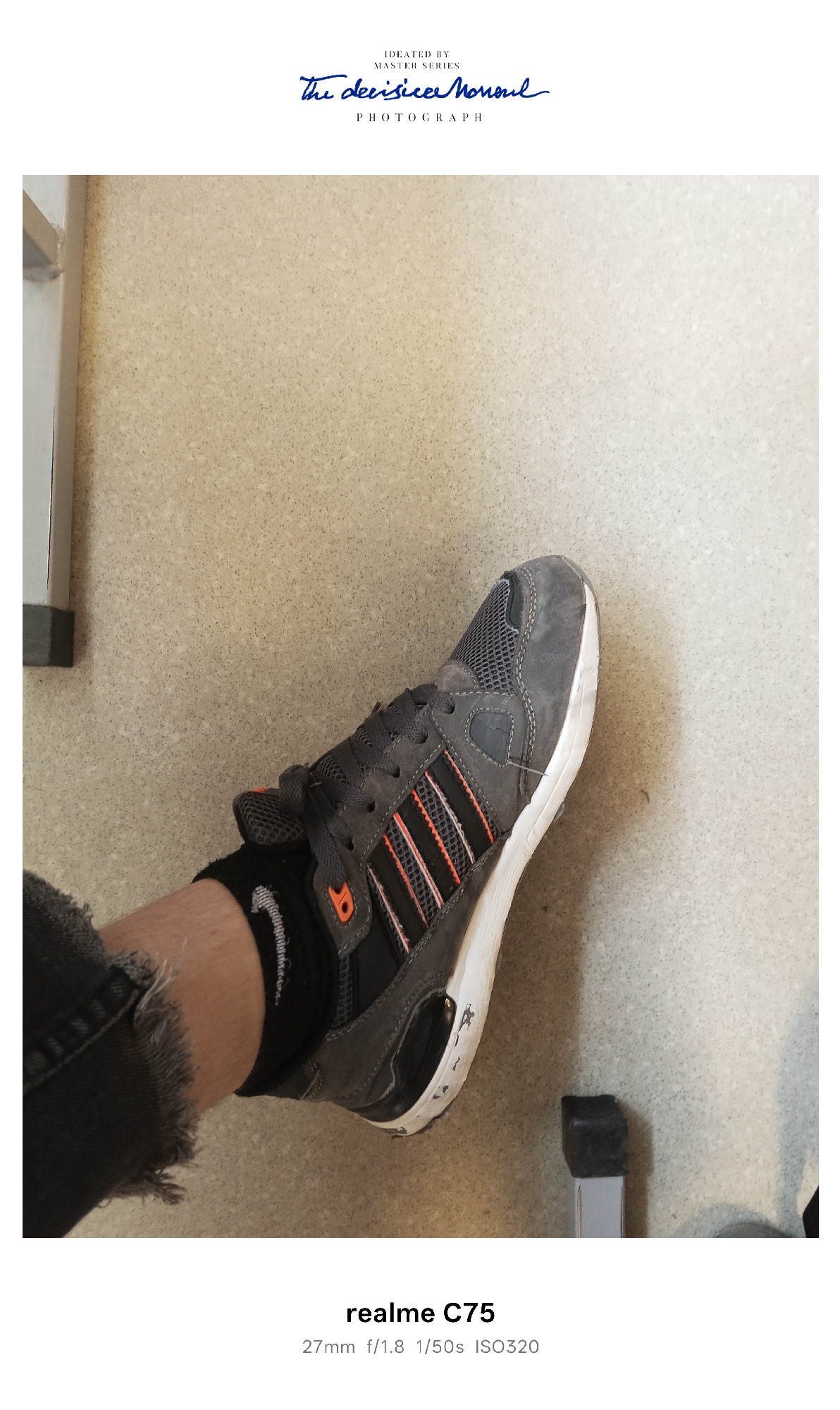 A close-up photograph taken indoors, possibly in Selty, Russia, shows a person's left foot wearing an Adidas running shoe with a black Nike sock. The shoe is gray with black and orange stripes and a white sole, appearing worn with scuff marks. The person is wearing distressed black denim jeans. The background features a mottled, neutral-toned wall and the legs of a metal chair. The image includes text indicating it was taken with a Realme C75 camera, with settings of 27mm, f/1.8, 1/50s, and ISO320. Additionally, text near the top of the image states "IDEATED BY MASTER SERIES The decisicee Moneul PHOTOGRAPH."