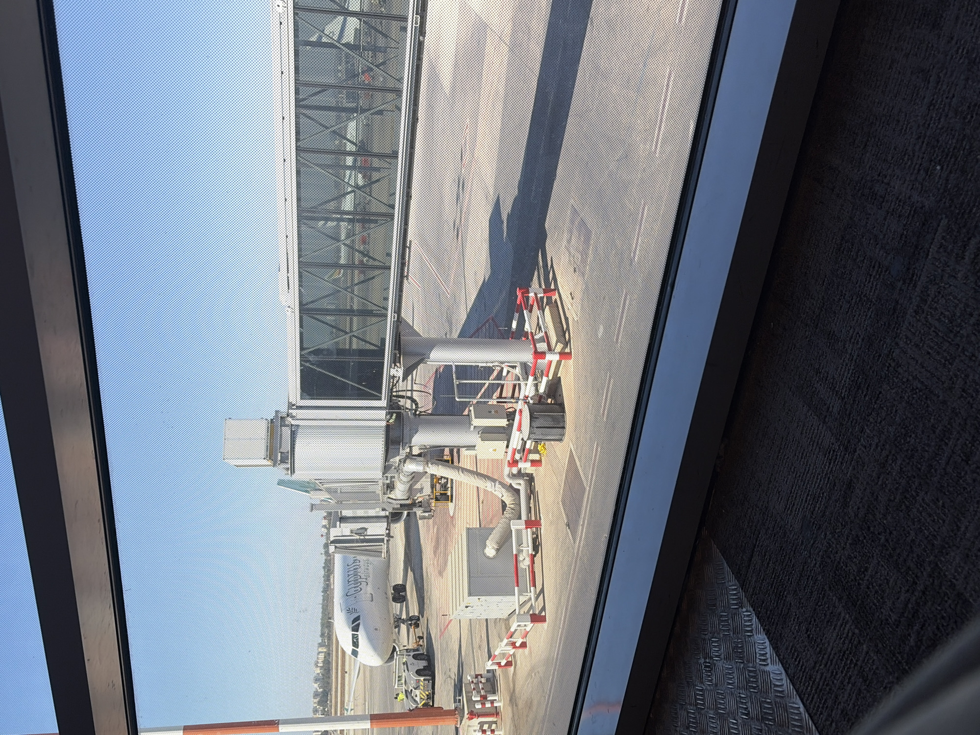 An airplane is parked at an airport gate in an exterior daylight shot. The airplane is white with a blue logo and text that reads "CYPRUS AIRWAYS". It is connected to a jet bridge, which is a covered walkway that transports passengers from the airport terminal to the aircraft. The jet bridge is an enclosed structure with windows, supported by columns. In the foreground, there is a carpeted floor and a metal floor plate. The background shows a city skyline and a clear blue sky. The location is the Ben Gurion Airport in Tel Aviv, Israel.