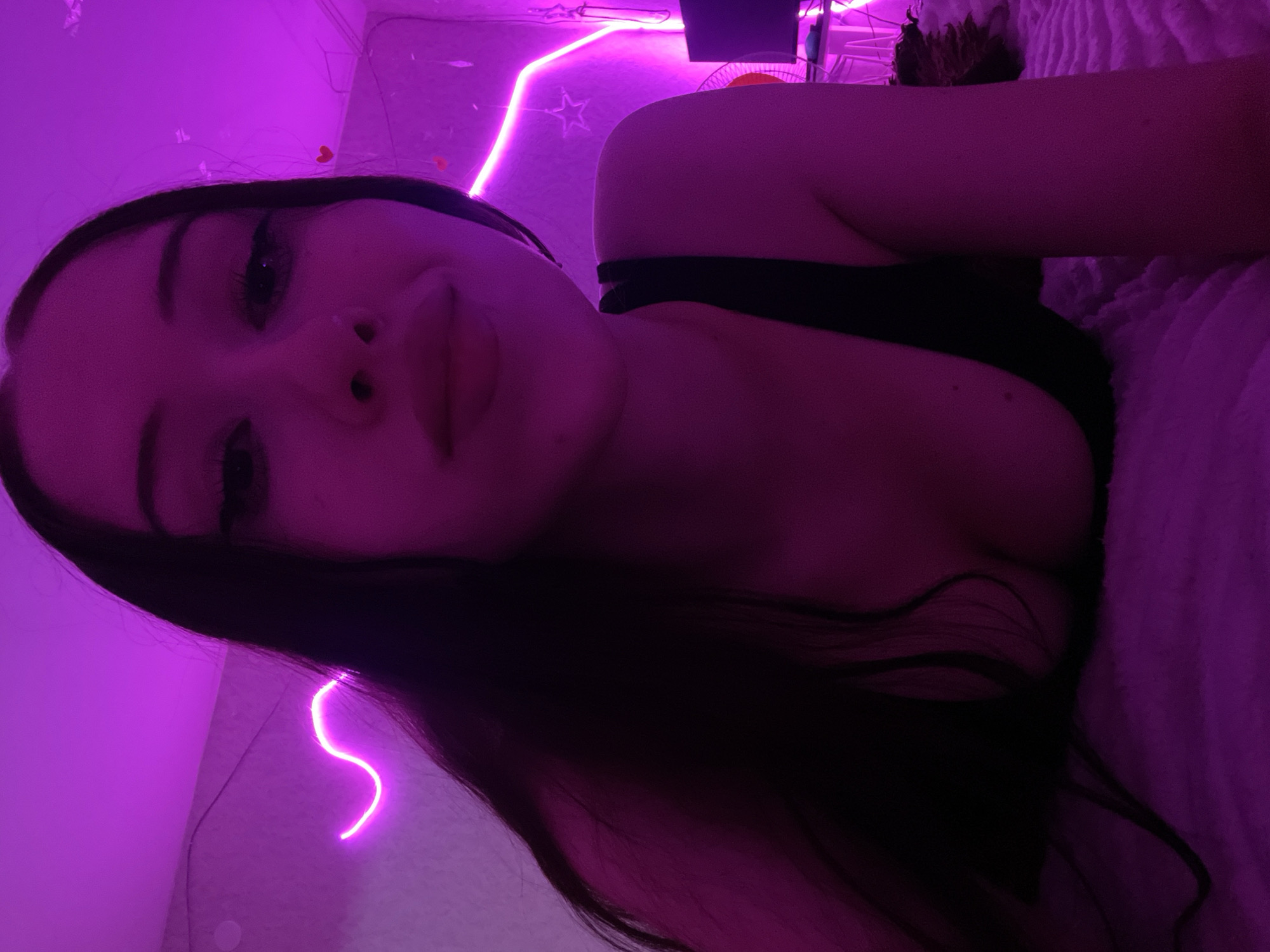 A young woman with long dark hair and makeup lies on a bed. She is wearing a black top. The room is lit with pink neon lights in a wave pattern and a star shape. A fan is visible in the background. The location is identified as Votkinsk, Russia.