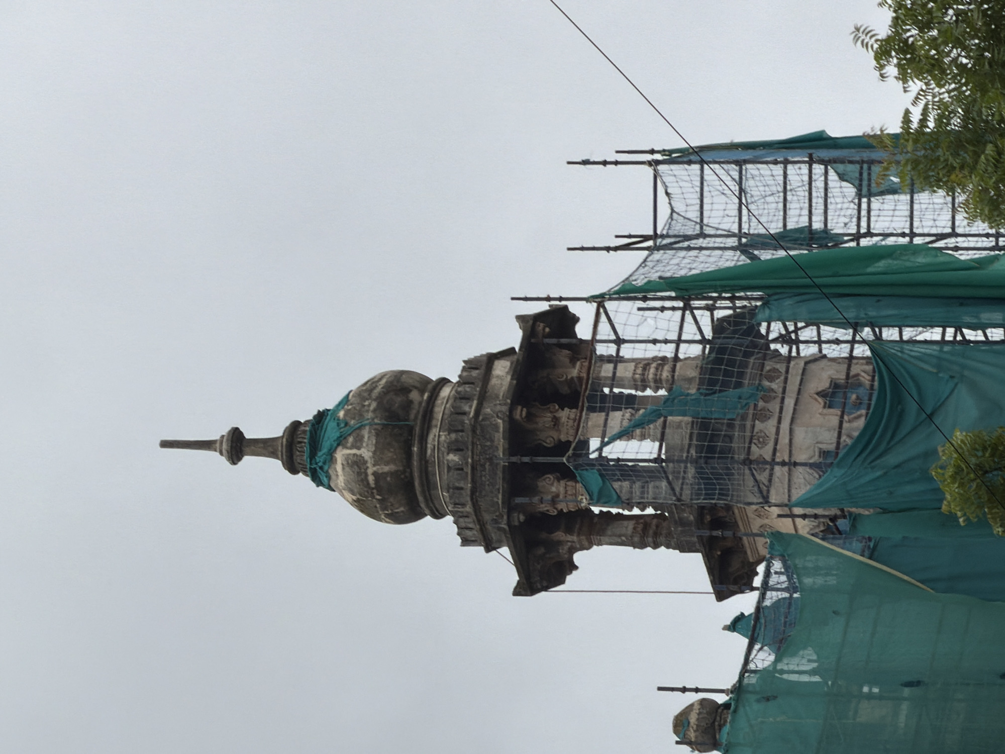 The image shows a close-up view of the minaret of a historical building, likely a mosque or tomb, in Mumbai, India. The minaret is under renovation, as evidenced by the scaffolding and green tarpaulins covering parts of the structure. The minaret has a domed top with a finial, and the stonework appears to be old and ornate, with carvings and decorative elements visible. The sky is overcast, suggesting a cloudy day. The scene is captured from a low angle, looking up at the minaret. The surrounding area appears to be a densely built-up urban environment, with trees visible in the foreground. The image provides a detailed view of the architectural style of the minaret and the ongoing restoration work.