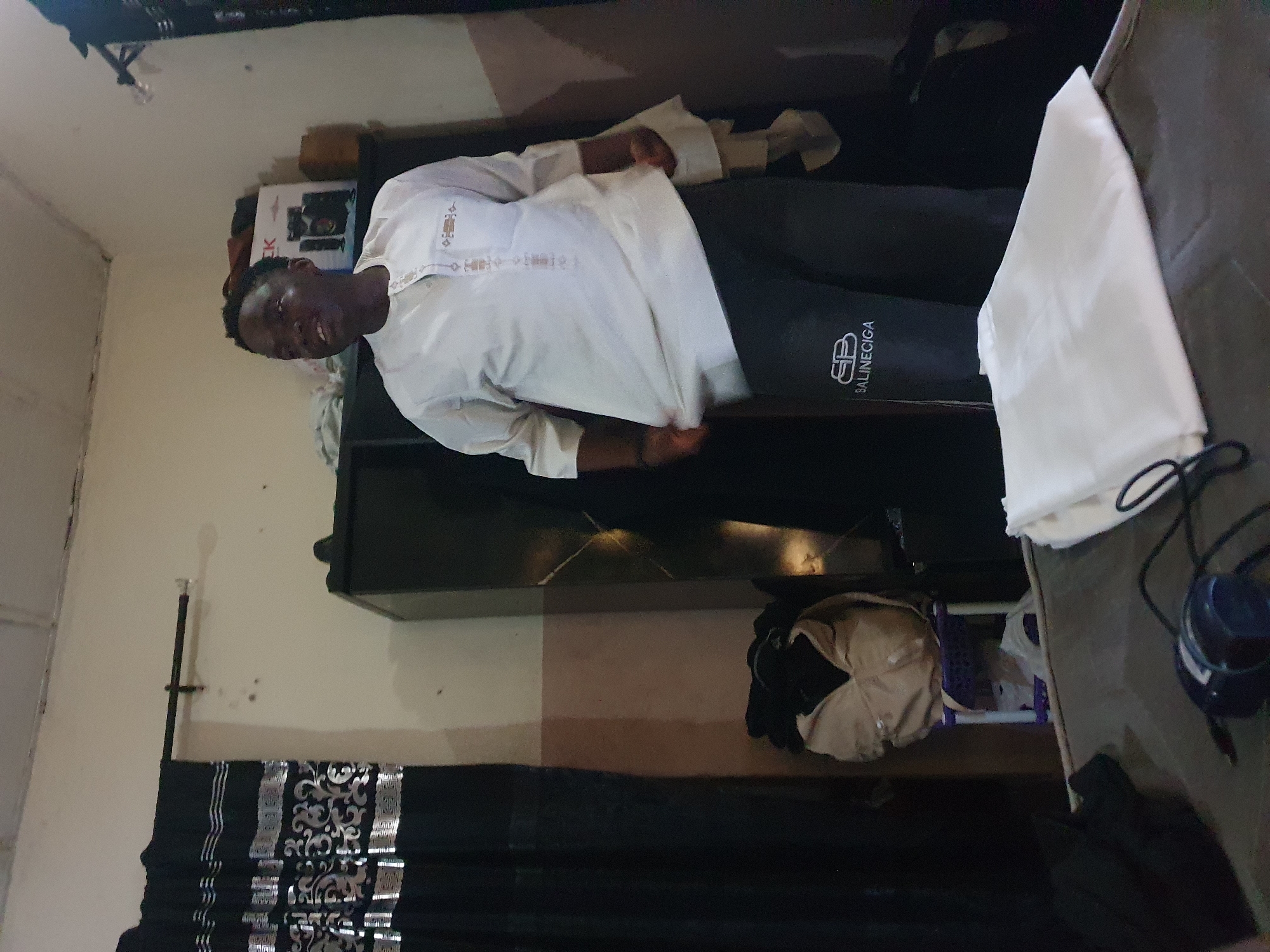 A man is in a room in Lokoja, Nigeria. He is wearing a white shirt and black pants with "BALINEICIGA" printed on the leg. There is a television, curtains, and a white bag in the room. He is standing in front of the television.