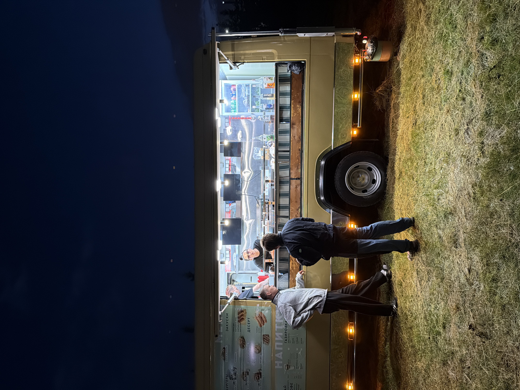An exterior, eye-level, vertical shot captures a food truck illuminated at night on a grassy area. Three individuals are visible. Two men stand in the foreground facing the food truck. One man, wearing a grey zip-up jacket and dark pants, faces the truck with his left hand resting on his hip. The other man, wearing a dark jacket and blue pants, faces the truck with his hands in his pockets. A third person, a woman with dark hair pulled back and wearing glasses and a black shirt, stands behind the counter of the food truck, appearing to be serving or interacting with customers. The food truck itself is tan with a lit menu board and display cases filled with bottles and drinks. Lights are on inside and underneath the truck, casting a warm glow on the surrounding grass. The sky above is a deep, dark blue, indicating nighttime. The overall scene suggests a casual outdoor dining or gathering at dusk or after dark.