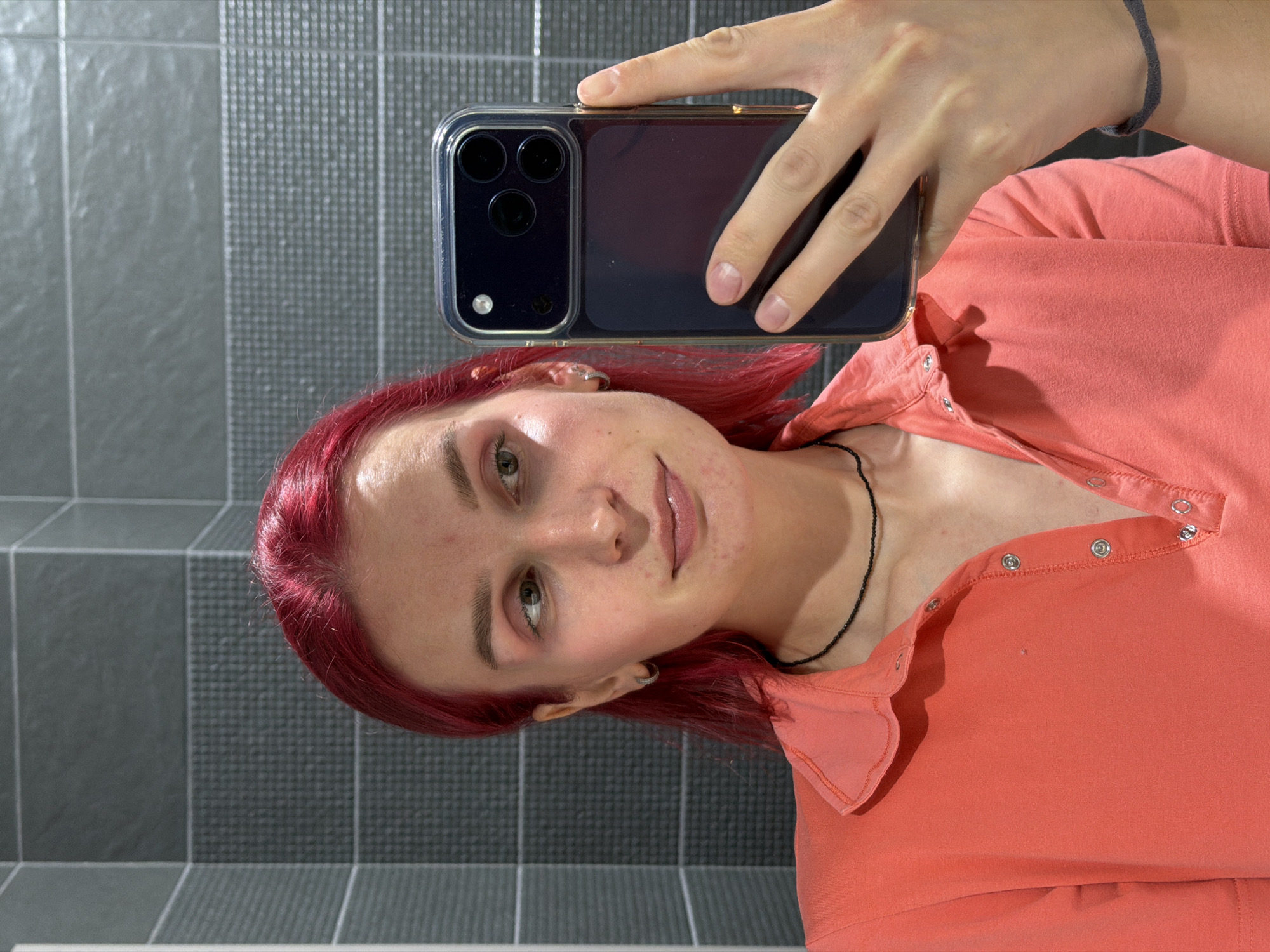 A female subject is depicted in a mirror selfie taken within a tiled interior space in Chaikovsky, Russia. The subject presents with shoulder-length, vivid red hair and a light complexion, exhibiting minor skin blemishes on her forehead, cheeks, and chin. She is observed looking towards her right, away from the camera's direct reflection. Her eyelids display a faint application of light pink eyeshadow. A small silver hoop earring is visible in her left ear. A thin black beaded choker necklace adorns her neck. She wears a coral-colored polo shirt, featuring a snap-button placket with several buttons unfastened. Her right hand, with natural, unpolished fingernails, holds a smartphone, identified as an iPhone 14 Pro or 15 Pro due to its triple-lens camera array and case design, positioned in the lower right foreground. A dark grey bracelet is present on her right wrist. The background consists of grey tiled walls, featuring smooth light grey rectangular tiles in the upper section, a middle band of darker grey textured tiles with a small grid pattern, and a lower band of plain dark grey tiles, indicating a modern interior environment.