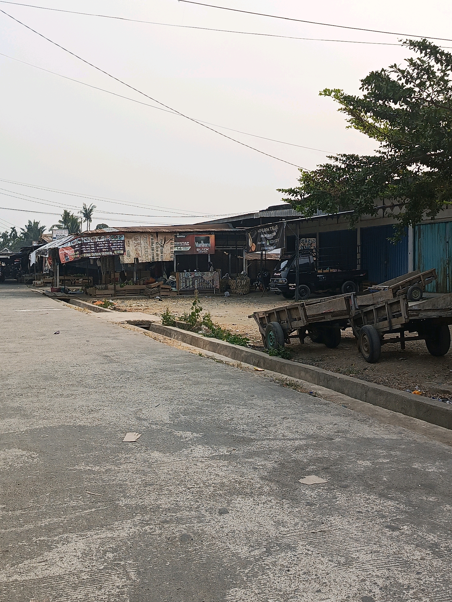 The image depicts a street scene in Megang Sakti I, Indonesia, likely in the morning or late afternoon, given the soft lighting. The foreground shows a paved road with a concrete curb and a drainage ditch. To the right of the road, there are several wooden carts with wheels, some appearing aged and weathered. Beyond the carts, a row of market stalls or small shops lines the street. These structures have corrugated metal roofs and are partially open, displaying various goods and signage, including what appears to be an advertisement for Marlboro cigarettes. A black pickup truck is parked in front of one of the stalls. In the background, palm trees and other vegetation are visible, along with utility wires strung across the sky. The overall impression is of a modest, functional marketplace in a tropical environment. There are no people clearly visible engaging in active transactions, though a few figures might be present within the stalls.