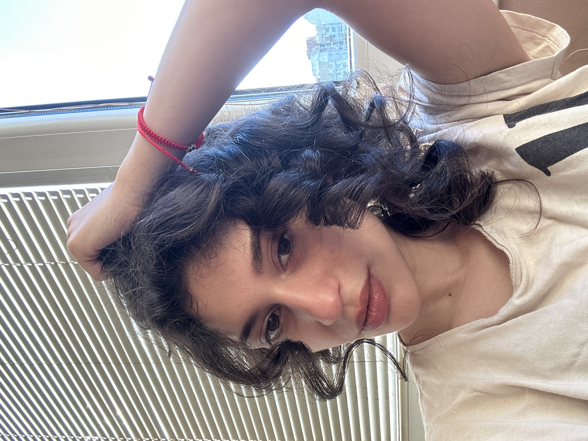 The image is a close-up, selfie-style shot of a young woman with dark, curly hair, wearing a light-colored t-shirt. She is indoors, with Venetian blinds visible in the background, suggesting a window. A bright red string bracelet is visible on her right wrist. The lighting suggests it is daytime. The setting does not provide sufficient information to determine a specific location within the city of Sofia, Bulgaria.
