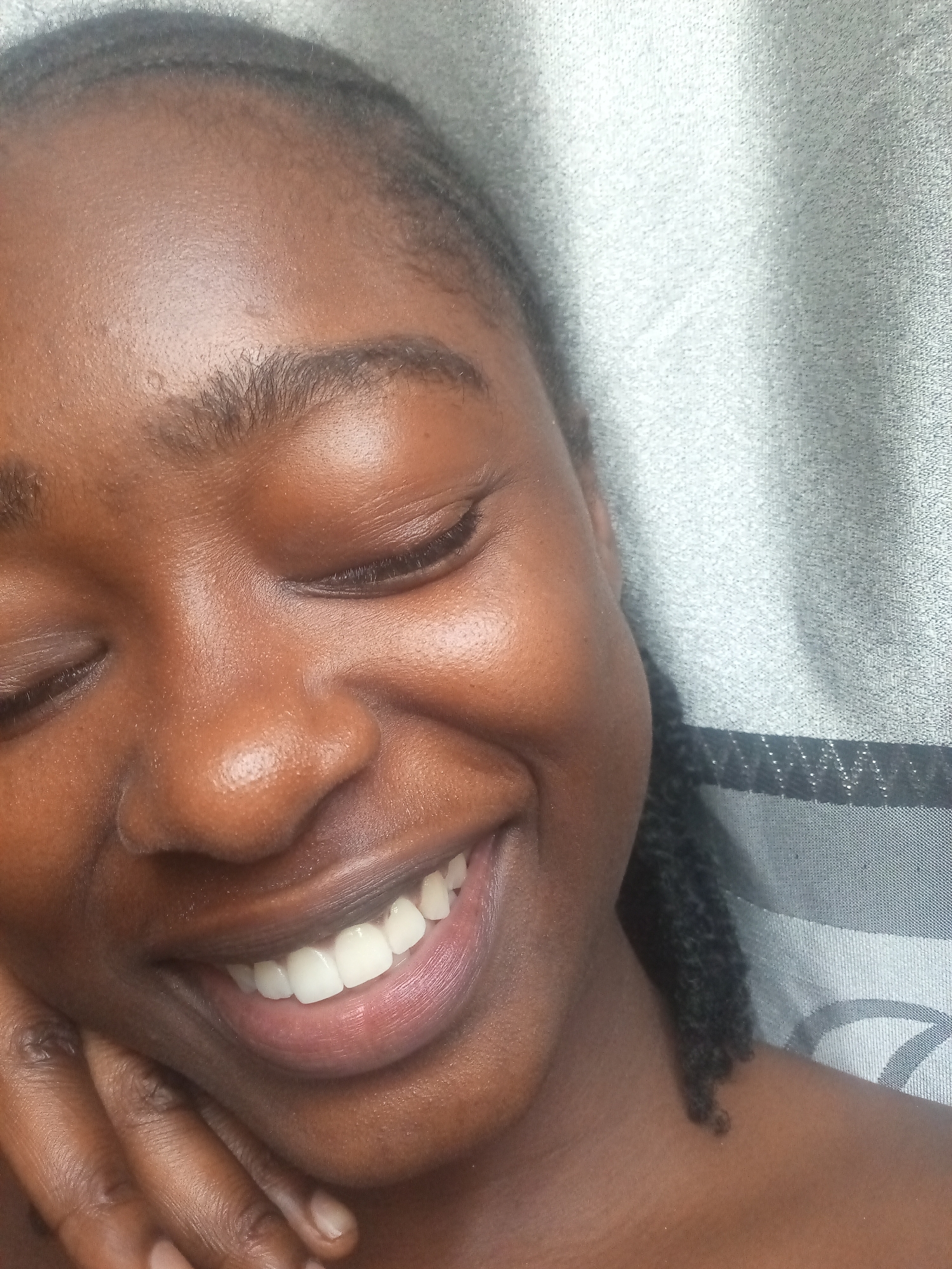 A close-up, head-and-shoulders shot captures a young woman's face from a slightly elevated angle as she smiles with her eyes closed. Her skin is a rich, dark brown and appears smooth and dewy. Her braided hair is pulled back, with a dark braid resting on her shoulder. The background consists of a textured, grey fabric with subtle metallic threads, possibly a curtain or upholstery. Her left hand is visible resting on her cheek, with her fingers gently cupping her jawline. Her teeth are bright white and evenly spaced. The lighting is soft, highlighting the contours of her face and the sheen on her skin. There are no discernible environmental clues to indicate a specific city or location.