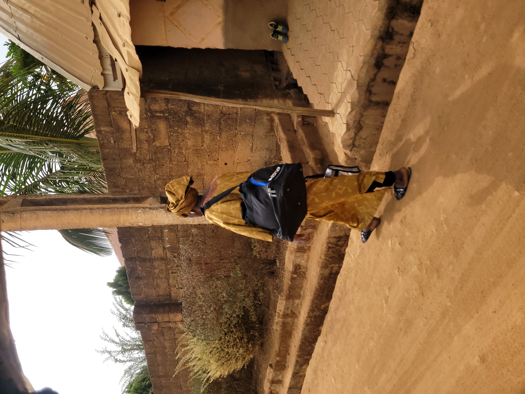 The image depicts an individual in Jos, Nigeria, walking on a path beside a building. The person wears a yellow garment, head wrap, and dark shoes. They carry a black sports bag over their shoulder. The building appears to be constructed from concrete bricks with a metal roof. The path is dirt. Vegetation grows along the base of the building. Another pair of shoes are located on the porch. The image appears to be taken during the day.