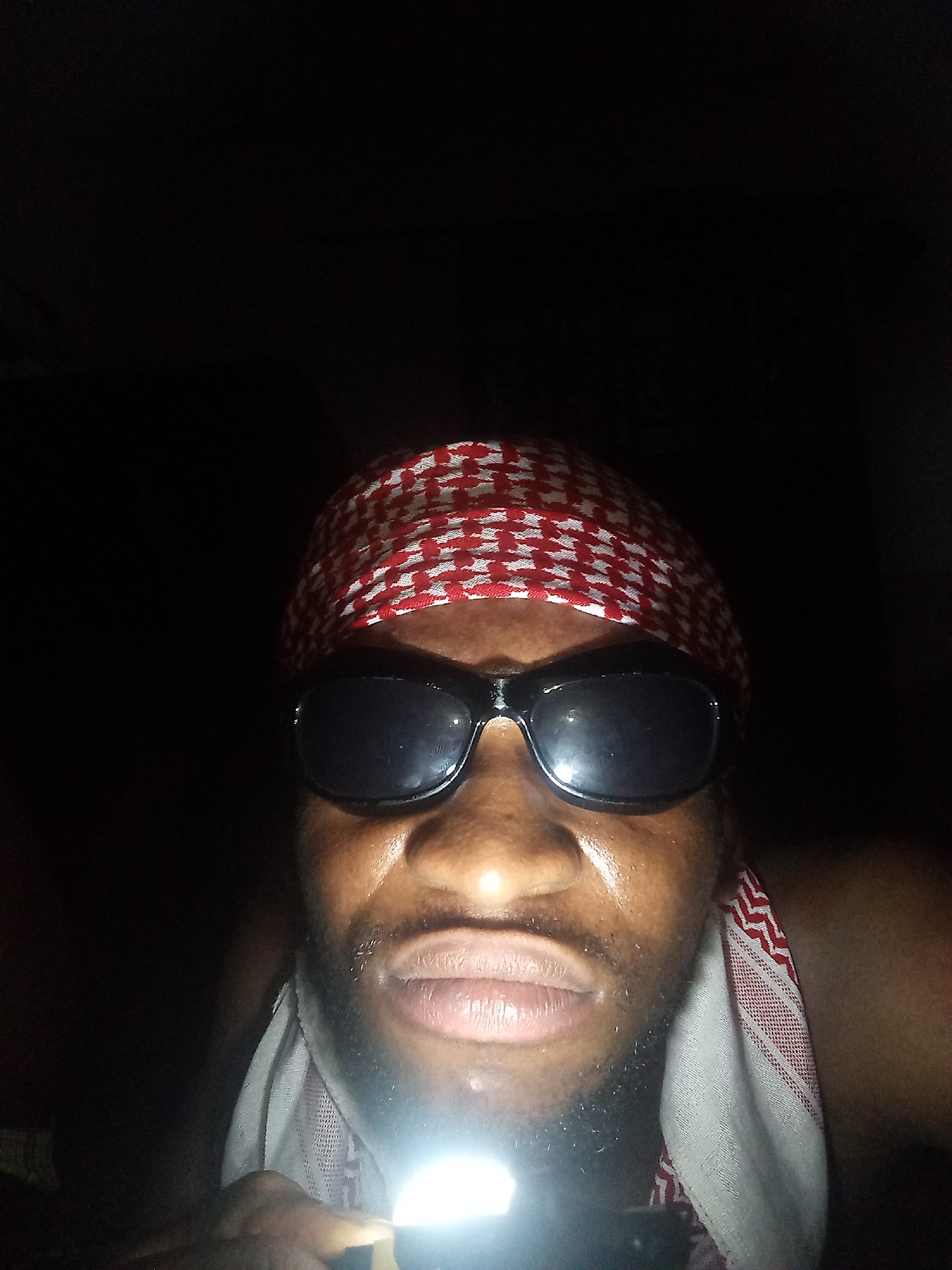 A close-up shot of a dark-skinned individual wearing a red and white patterned headscarf and dark sunglasses. The individual is illuminated by a light source held below their face, which is the primary source of light in the image. The background is dark and indistinct. Location not discernible from the image.