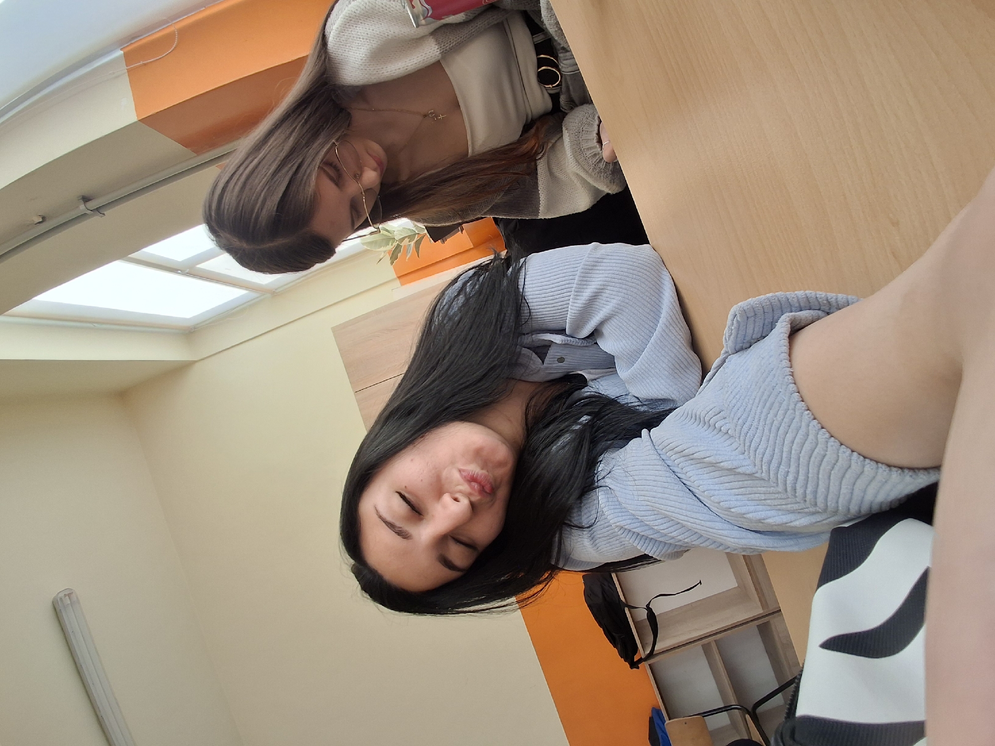 Two young women are captured in a casual, indoor setting, likely in Sofia, Bulgaria, as indicated by the location context. The image is taken from a slightly elevated, rotated perspective, looking down towards the women who appear to be seated or leaning at a wooden desk.
The woman in the foreground has long, dark hair and is wearing a light blue, ribbed long-sleeved top. Her eyes are closed, and her lips are pursed in a playful expression, possibly blowing a kiss or pouting.
Behind her, another woman with long brown hair is partially visible. She is wearing eyeglasses and what appears to be a light-colored tank top under a grey and white striped or textured cardigan. A delicate gold-colored necklace is visible around her neck, and she has hoop earrings. Her gaze is directed downwards, suggesting she might be looking at something on the desk or simply lost in thought. A small potted plant can be seen behind her.
The setting features a prominent wooden desk surface that occupies a significant part of the frame. The background shows light cream-colored walls, with an orange accent wall or paneling section. Above, several rectangular skylights or large ceiling windows are visible, indicating abundant natural light, suggesting it is daytime. Some shelving units and a black bag are faintly seen in the background. No visible text is present in the image. The overall mood appears relaxed and informal.