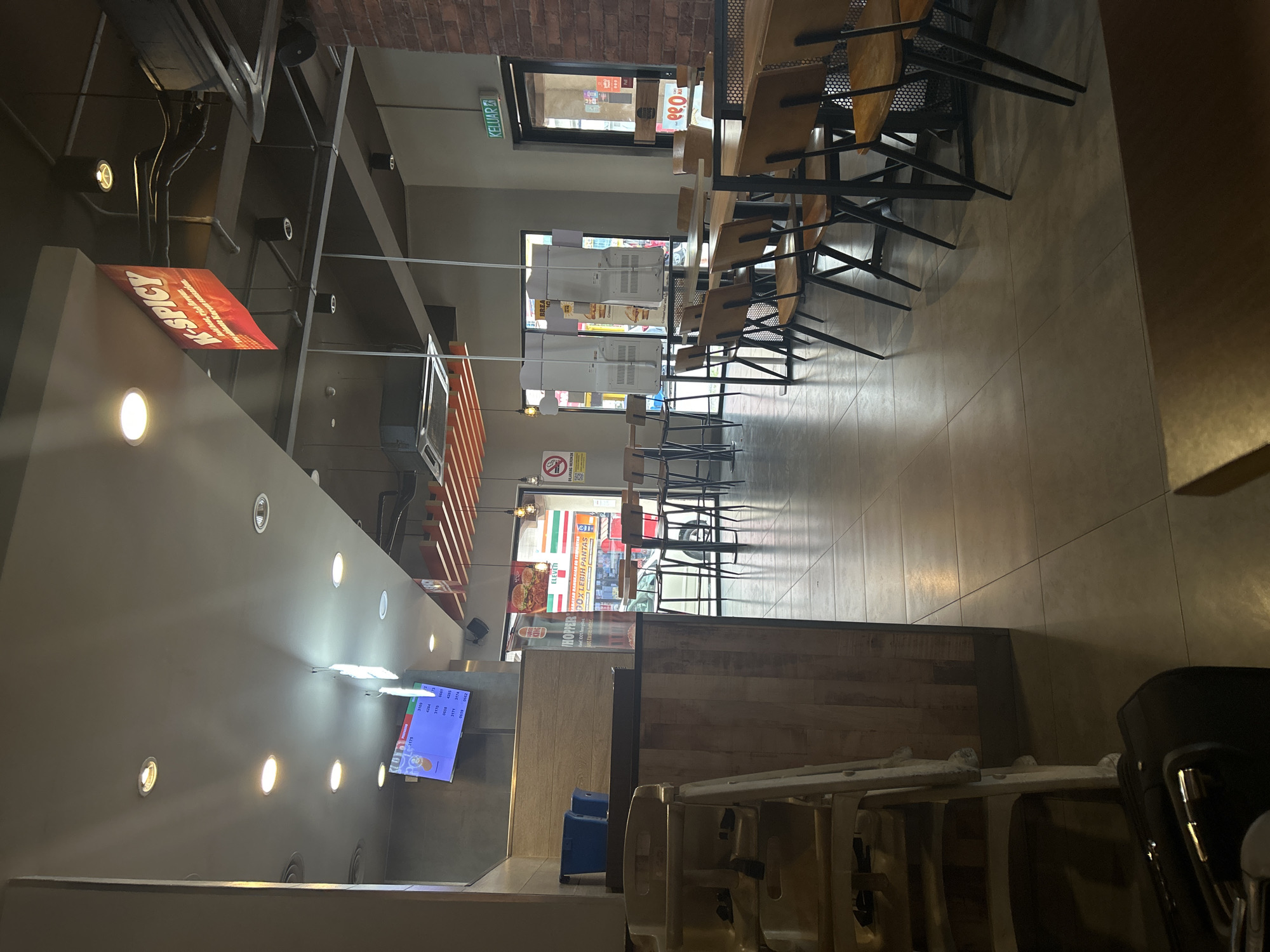 Here is a description of the image:
The image is taken inside a restaurant, possibly a Burger King, located near a 7-Eleven in Subang Jaya, Malaysia. The restaurant is empty with rows of tables and chairs neatly arranged, suggesting it's either early in the day or after closing hours. A sign "K-SPICY" is visible above the ordering counter. There is a TV screen displaying numbers behind the counter, likely used for order tracking. A "KELUAR" (Exit) sign is visible above a door. A "DILARANG MEROKOK" (No Smoking) sign is posted, indicating regulations within the area. A couple of blue luggage trolleys are located near the counter.