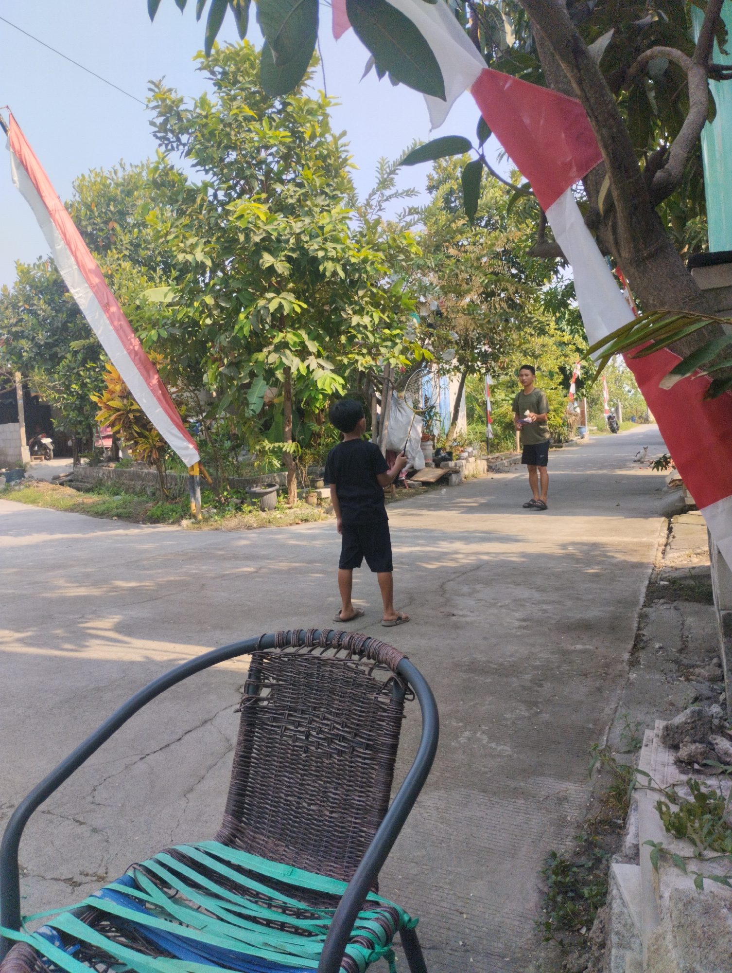 In the city of Karangraharja, Indonesia, a street scene unfolds under a clear sky, framed by trees adorned with red and white Indonesian flags. In the foreground, a woven chair with a green and blue striped seat faces away from the camera, suggesting it's unoccupied or has just been vacated. Two young males are present on the paved street. One, wearing a black t-shirt and shorts, stands facing away from the camera, holding a badminton racket. Further down the street, another young male in a green t-shirt and dark shorts stands, holding an object. The street is lined with houses and vegetation, indicating a residential area. A white cat is visible on the right side of the street in the mid-ground. The overall atmosphere is one of a casual outdoor activity in a village or suburban setting.