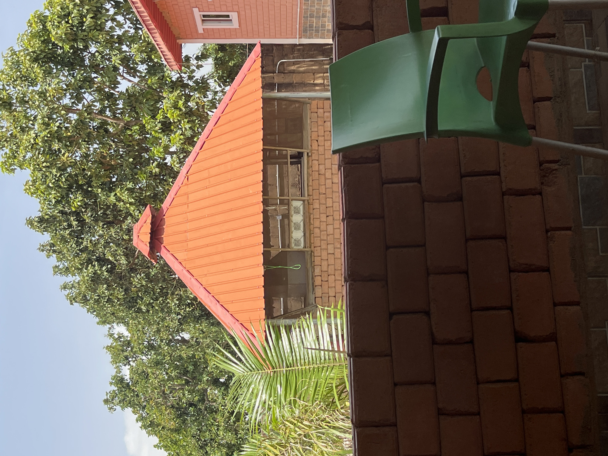 A view of a building with a red corrugated roof and brick walls. In front of the building is a green plastic chair. The building is partially obscured by a brick wall and lush green trees. The scene appears to be outdoors, under a bright, partly cloudy sky. The overall impression is one of a casual outdoor space, possibly a restaurant or a guesthouse, in a tropical or subtropical climate. The architectural style and vegetation suggest it could be located in Jos, Nigeria.
