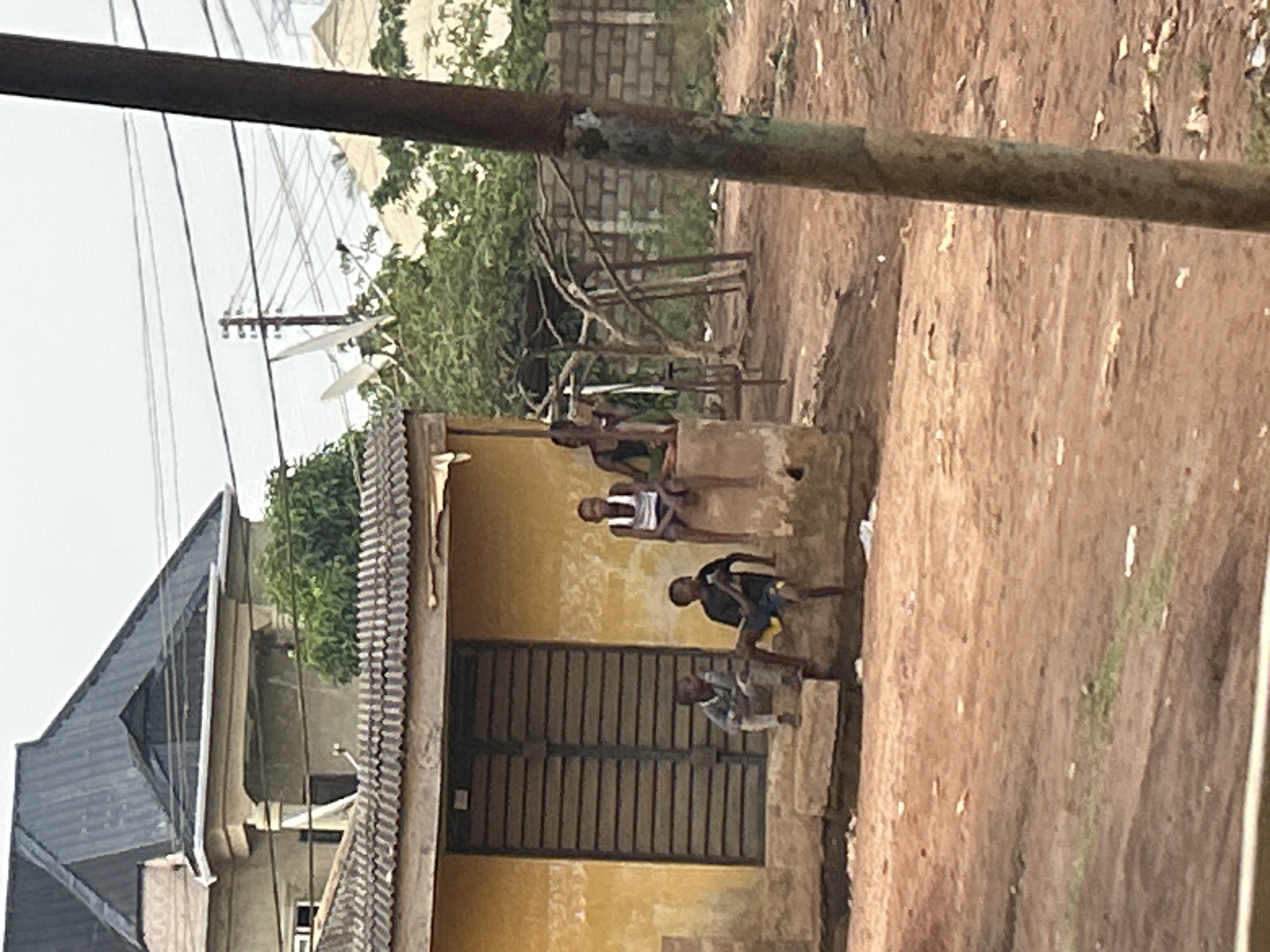 The image, oriented vertically, captures an outdoor scene in what appears to be a community setting.
**Subjects:** Several young individuals, likely boys, are visible. Three are prominent: one in a white tank top and dark shorts is sitting on a concrete platform; another, wearing a dark top and light shorts, is on a lower step; and a third, in a light top and dark shorts, is positioned near a large, closed metal gate. Other figures are partially obscured further back.
**Objects:** A single-story building with yellow-painted walls and a dark, corrugated metal roof occupies the left side. The large, dark, corrugated metal gate is part of this building. A concrete structure in front of the building serves as a seating area. A dense network of overhead electrical wires and utility poles is visible against the light sky. Two small satellite dishes are attached to one of these poles. A thick, dark, metallic object, possibly a pipe or beam, runs diagonally across the upper portion of the frame. The ground is unpaved, characterized by reddish-brown dirt. Green foliage from trees and bushes is scattered in the background. Other buildings with dark, angled roofs are partially visible in the distance.
**Scene/Setting:** The scene depicts a casual outdoor gathering in a residential or semi-urban area. The unpaved ground, visible utility infrastructure, and building architecture suggest a locale in a developing region.
**Activity or event taking place:** The individuals appear to be resting or socializing outside the building. They are in relaxed postures, suggesting a common daily scene rather than a specific event.
**Notable details:** The time of day is daytime, under an overcast sky, indicated by the bright, diffused light and absence of harsh shadows. The weather appears calm. There is no visible text in the image.