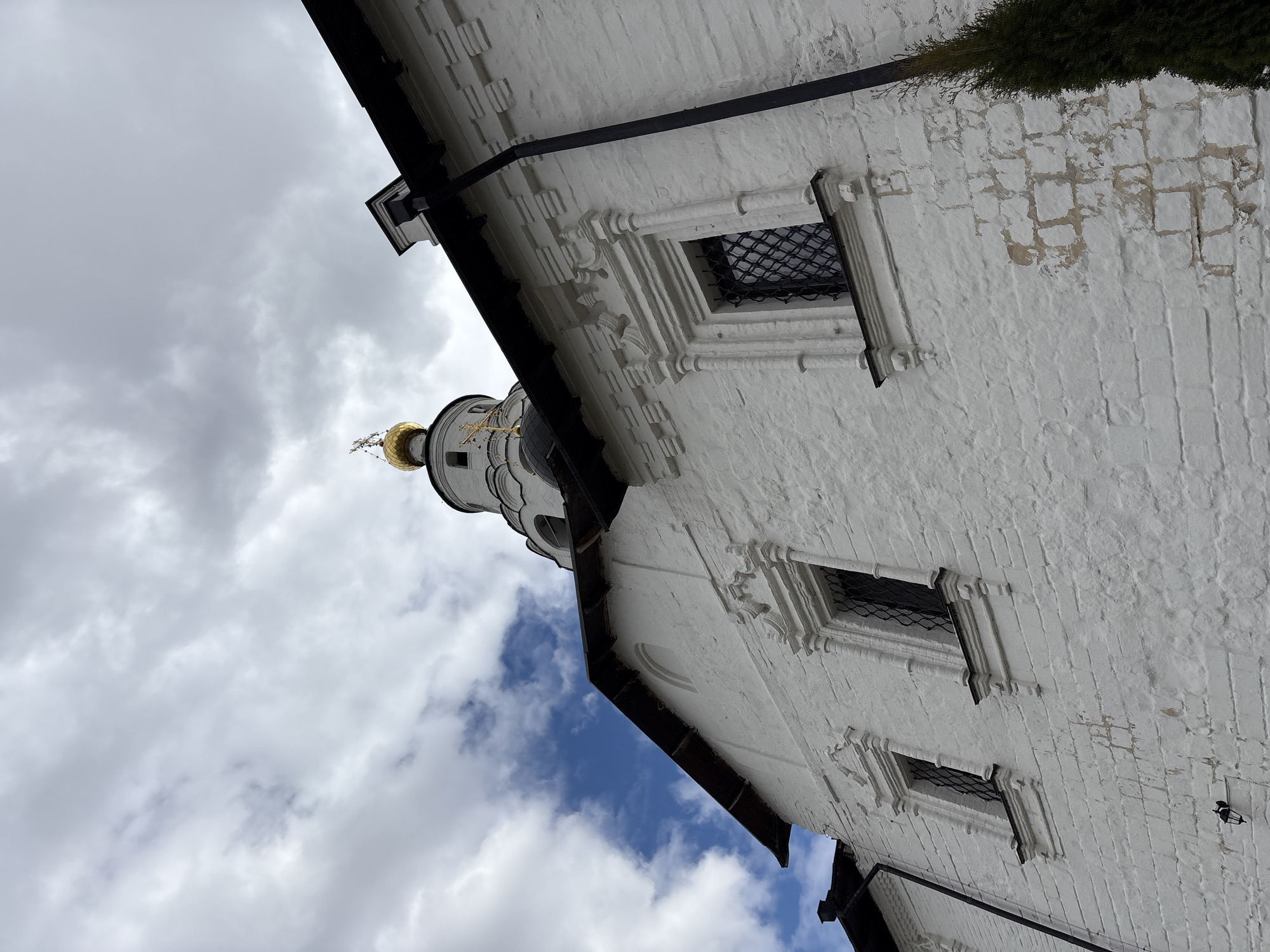 An upward, angled view of a white brick building with architectural details, including ornate window frames and a dark trim running along the roofline, and a golden domed tower. The sky is visible in the background, with a mix of white clouds and patches of blue. The structure appears to be part of a larger religious complex, likely a church or monastery. The location is Sviyazhsk, Russia.