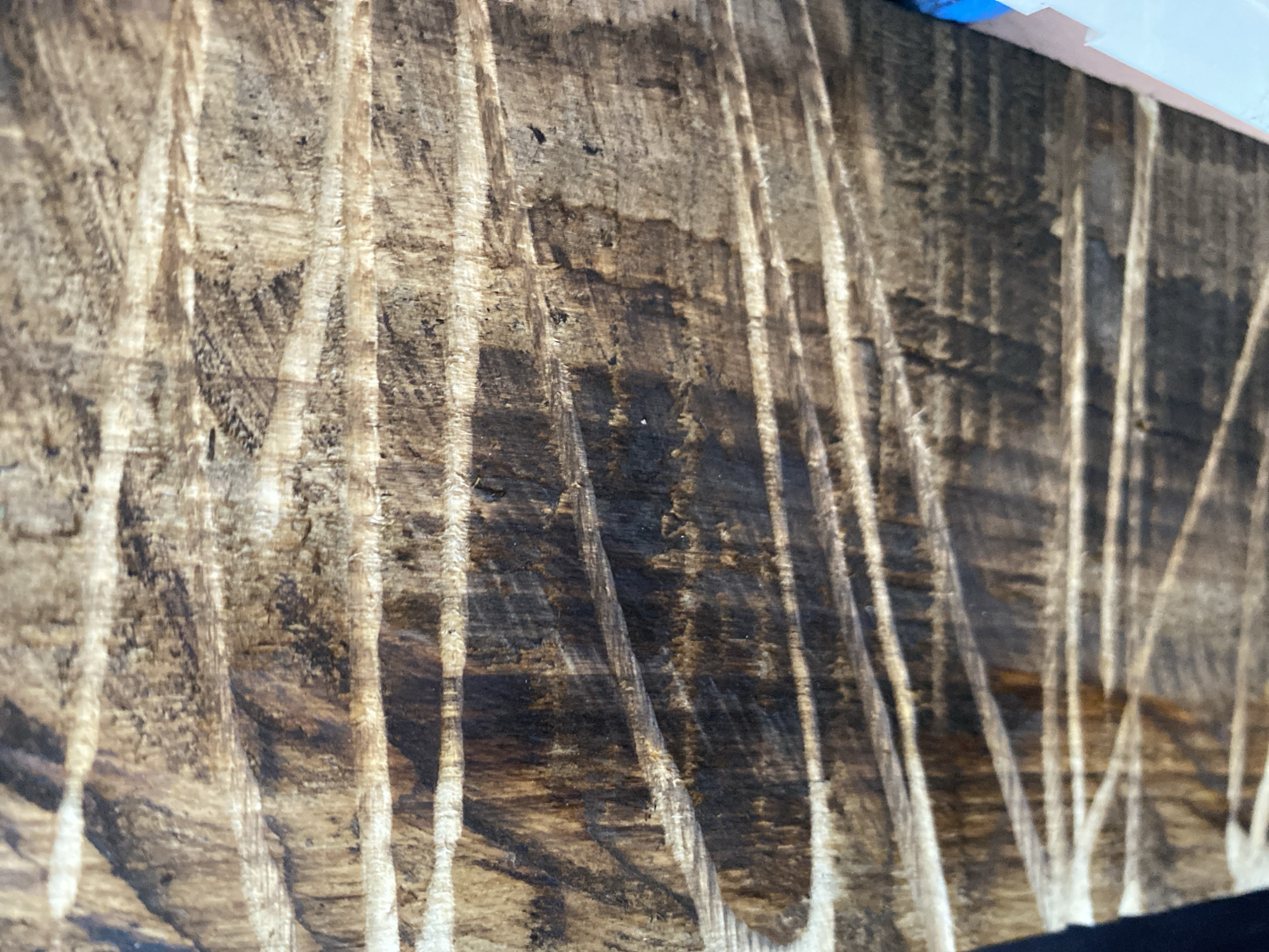 The image is a close-up, angled shot of a weathered wooden surface, possibly a fence or a section of a building, exhibiting distinct grain patterns and horizontal scoring or carving marks. The wood is dark brown with lighter, textured streaks, suggesting age and exposure to the elements. The lighting creates shadows that emphasize the depth and texture of the wood. The background is not clearly visible, but hints of light blue and white material appear at the bottom right edge, possibly part of an adjacent structure or object. No people or specific objects are identifiable within the wooden surface itself. The image does not contain any contextual information about its location in Kisliakovskaya, Russia.
