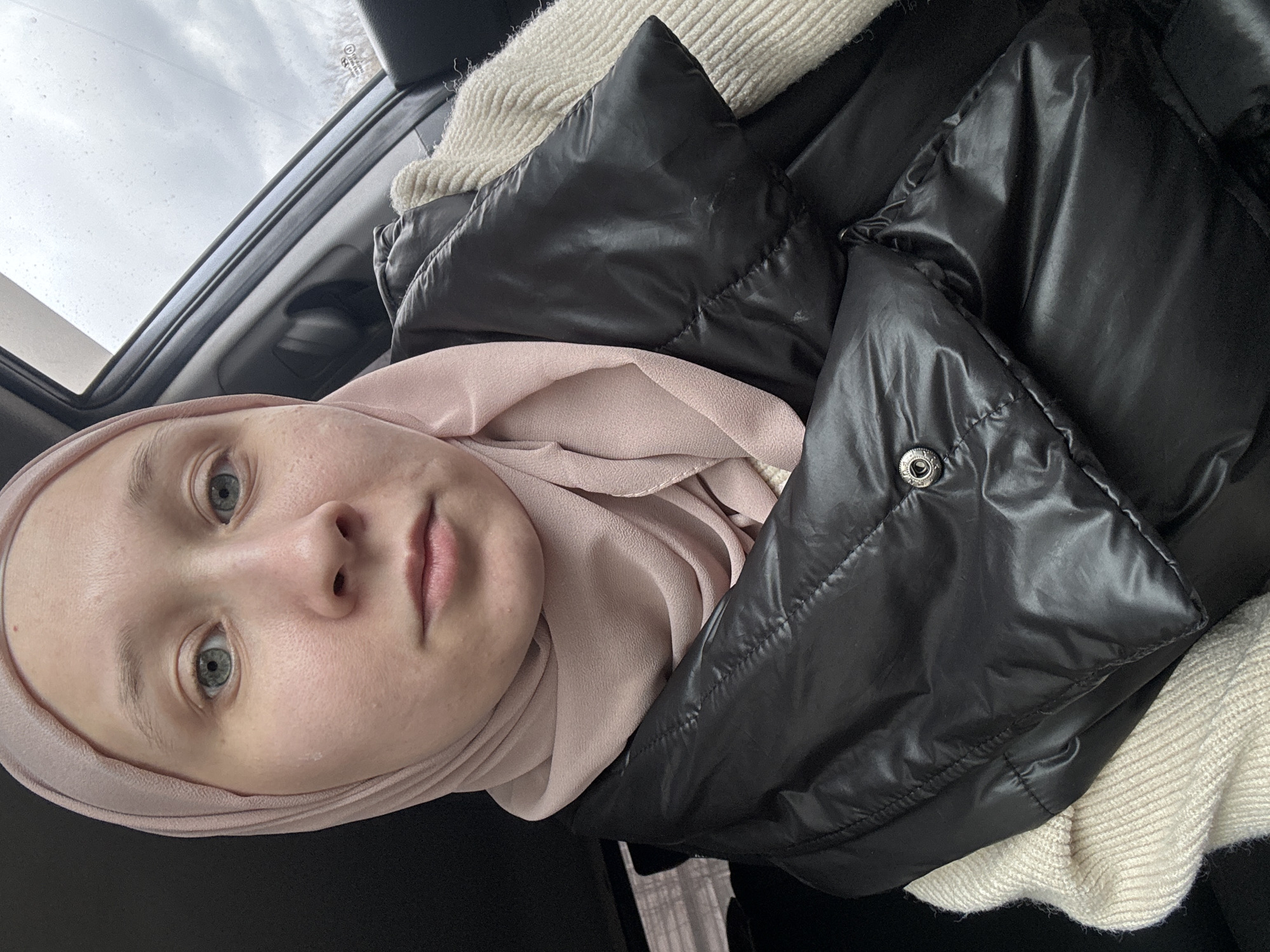A young person is captured in a close-up portrait, likely a selfie, inside what appears to be a car. The individual, who has fair skin and light blue-green eyes, looks directly at the camera with a calm, neutral expression. They are wearing a light pink hijab neatly wrapped around their head.
Over the hijab, they wear a black, shiny puffer jacket, which has visible stitching creating quilted sections. Beneath the puffer jacket, a cream-colored, chunky knit sweater is visible, providing a contrasting texture. A silver-colored snap button is present on the front of the puffer jacket.
The setting appears to be the interior of a vehicle, suggested by the car window frame visible in the upper part of the image. Through the window, an overcast sky is seen, with visible raindrops or water stains on the glass, indicating a dull or wet day. Textual markings are discernible on the car window, including "FUYAO", "TOUGHENED", "ECE R43", "DOT", and "AS2", which are typical automotive glass manufacturing and safety codes.
The activity taking place is a still portrait, possibly self-recorded due to the close-up, head-on angle. The scene depicts a moment of quiet observation from within a vehicle in Chaikovsky, Russia, on an overcast day.