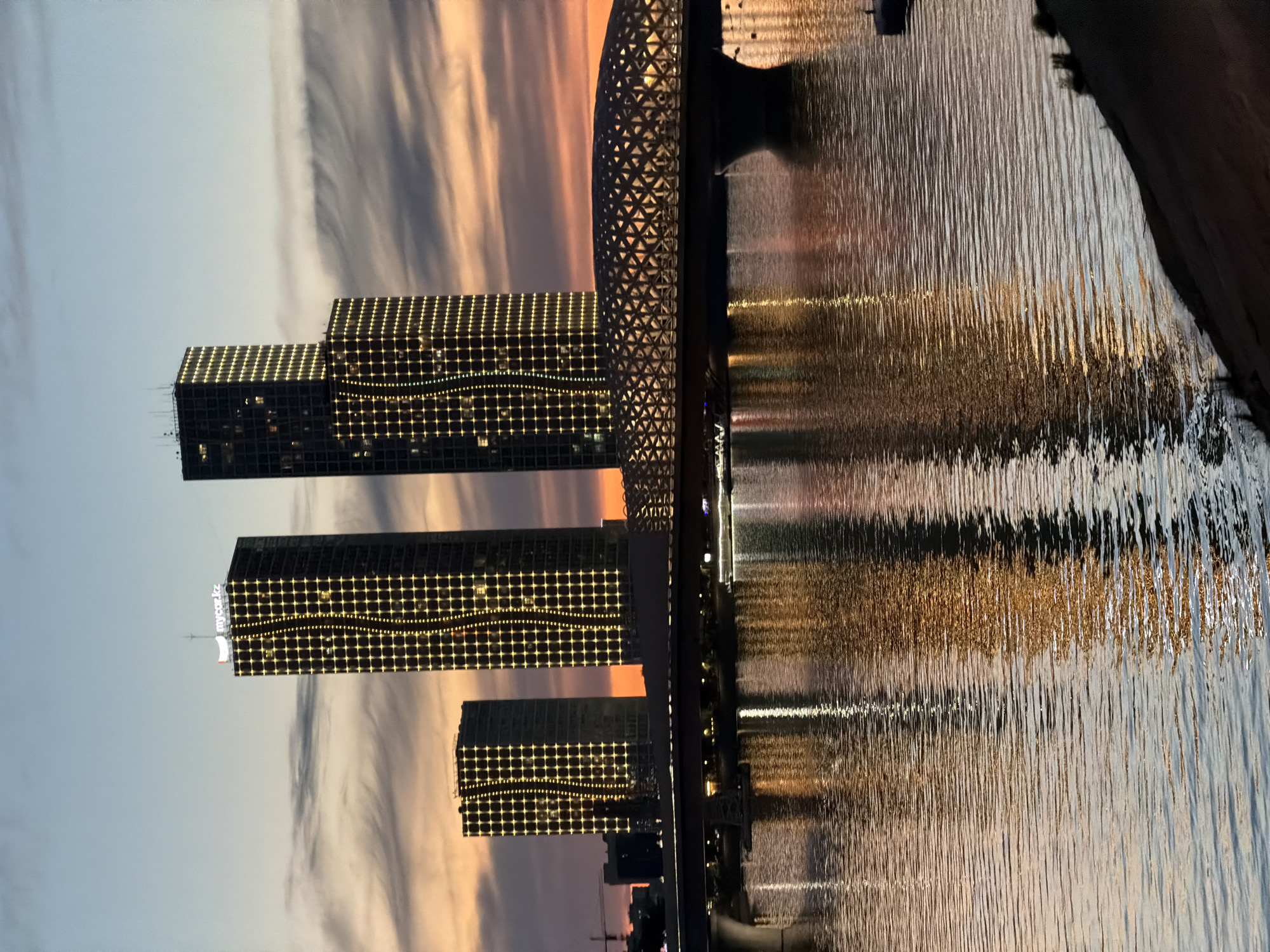 The image captures a twilight scene of the city of Astana, Kazakhstan, specifically focusing on the futuristic architecture along the Ishim River. In the background, a cluster of modern skyscrapers dominates the skyline. The tallest buildings feature intricate grid patterns illuminated by warm yellow lights, creating a striking visual effect against the darkening sky. One of the buildings displays the white text "mycar.kz" at its apex. A uniquely designed bridge, with a geometric, lattice-like structure and illuminated edges, spans the river in the foreground, connecting the cityscape. The river itself reflects the lights of the buildings and the bridge, creating shimmering patterns on its surface. The sky displays a gradient of colors, transitioning from a pale blue at the top to warmer hues of orange and pink near the horizon, indicative of a sunset or sunrise. The overall impression is one of a modern, vibrant urban landscape set against a dramatic natural backdrop.