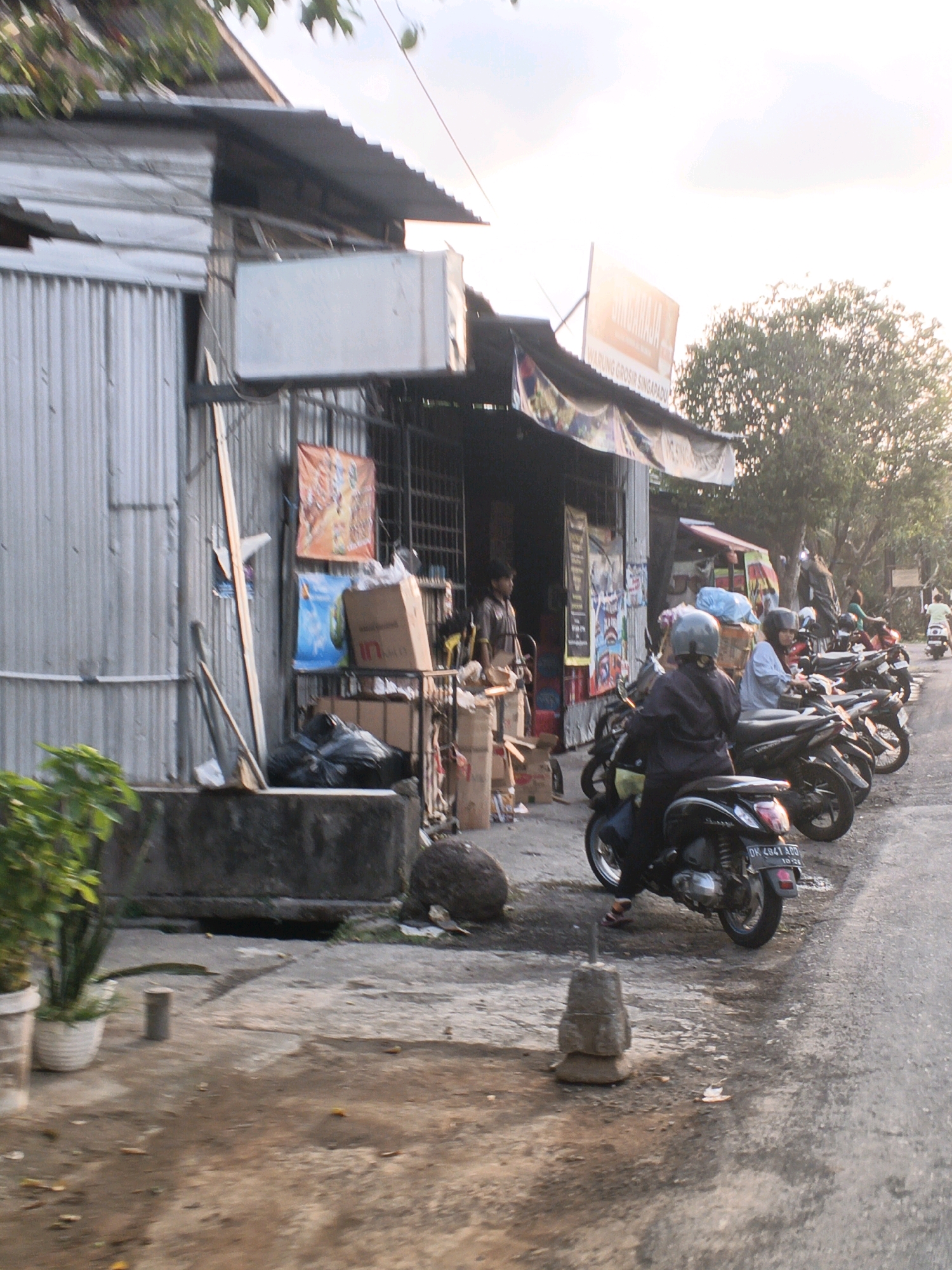 The image depicts a street scene in Sukawati, Indonesia, likely in the late afternoon. A corrugated metal building, appearing to be a shop or market stall, dominates the left side of the frame. Various items are displayed outside, including posters, boxes, and bags of trash. A person is visible inside the building through an open doorway, and another person stands to the left of the building. Several motorbikes are parked or moving along the road in front of the building. Some of these motorbikes have riders, many wearing helmets. A woman in a grey helmet and dark jacket is on a motorbike in the foreground, and another person in a light blue shirt and helmet is on a motorbike behind her. A person in a light brown shirt and helmet is seated on a motorbike further back. In the background, trees and a bright sky suggest an outdoor environment. The ground is paved with asphalt and appears somewhat wet or dirty. The overall impression is one of a busy commercial area with activity centered around motorbikes and local businesses.